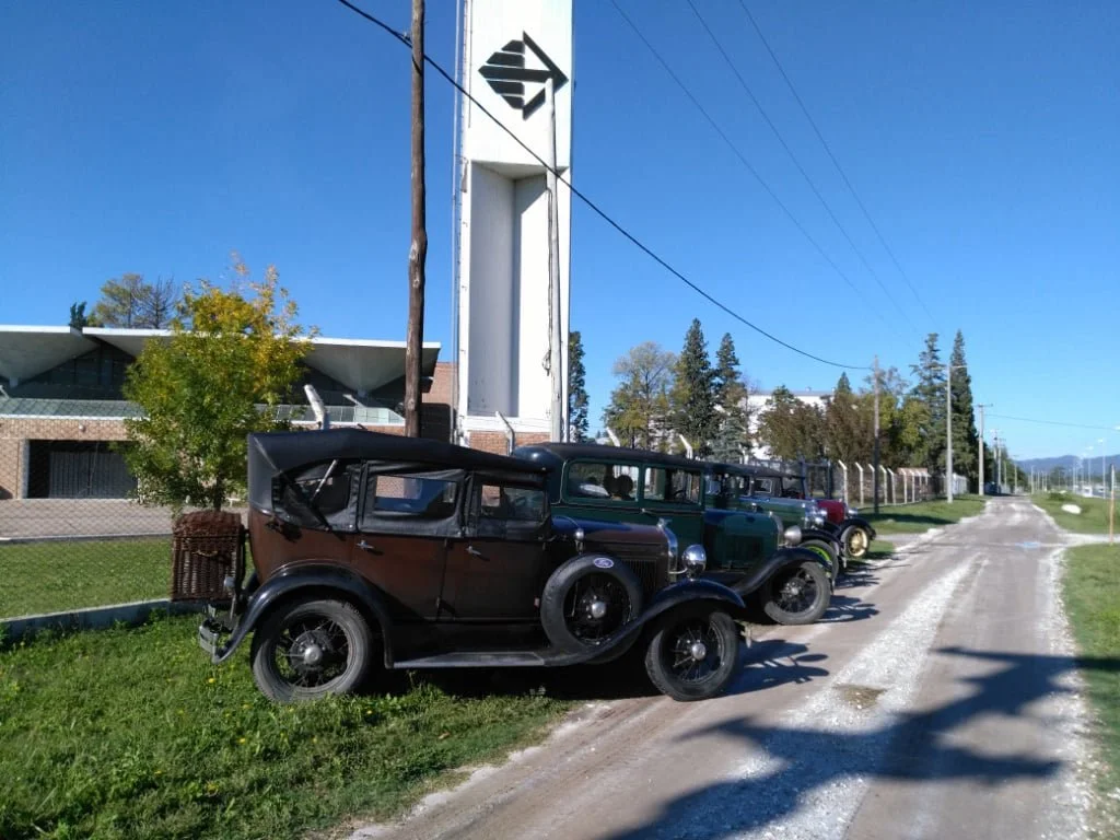 Cuatro autos clásicos estacionados en una calle de tierra junto a una cerca, con un edificio y un cartel alto con logotipo en el fondo, bajo un cielo despejado.