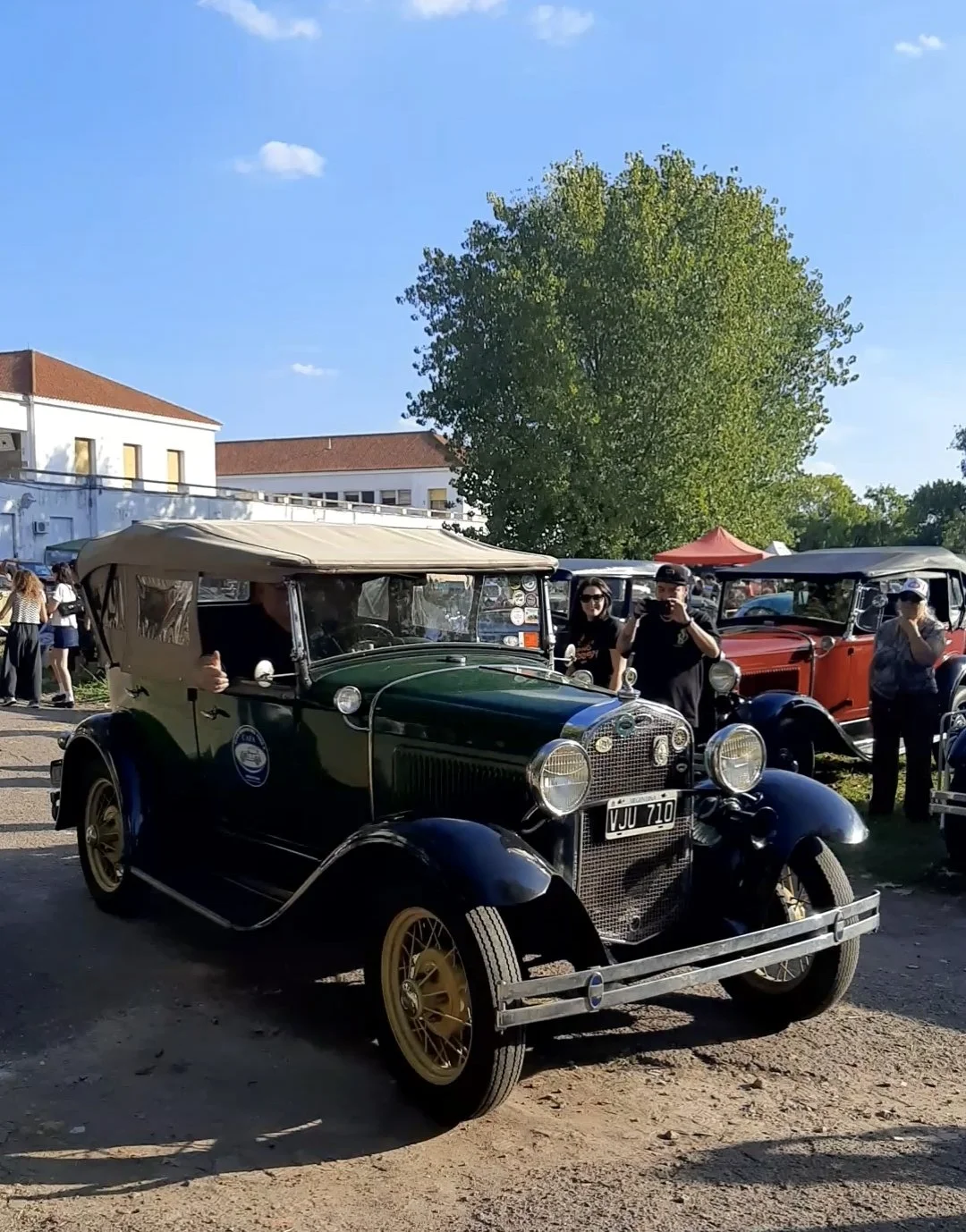 Coche vintage de color verde y negro en un evento al aire libre con varias personas y otros autos antiguos en el fondo, bajo un cielo despejado.