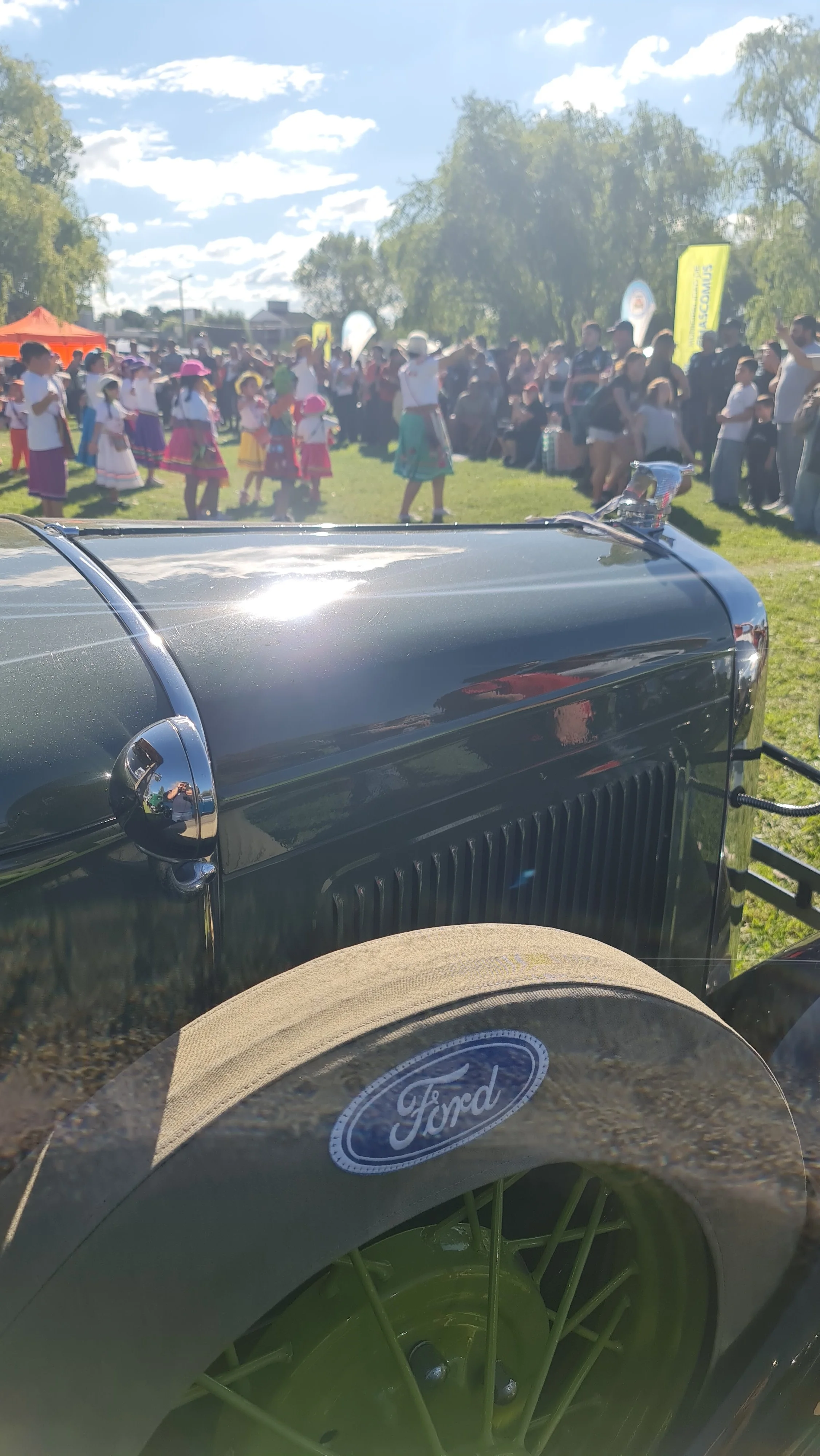 Evento con muchas personas, niños con trajes tradicionales, banderas y carpas, en un día soleado, vista de cerca de un vehículo antiguo y un sombrero con logotipo de Ford en primer plano.