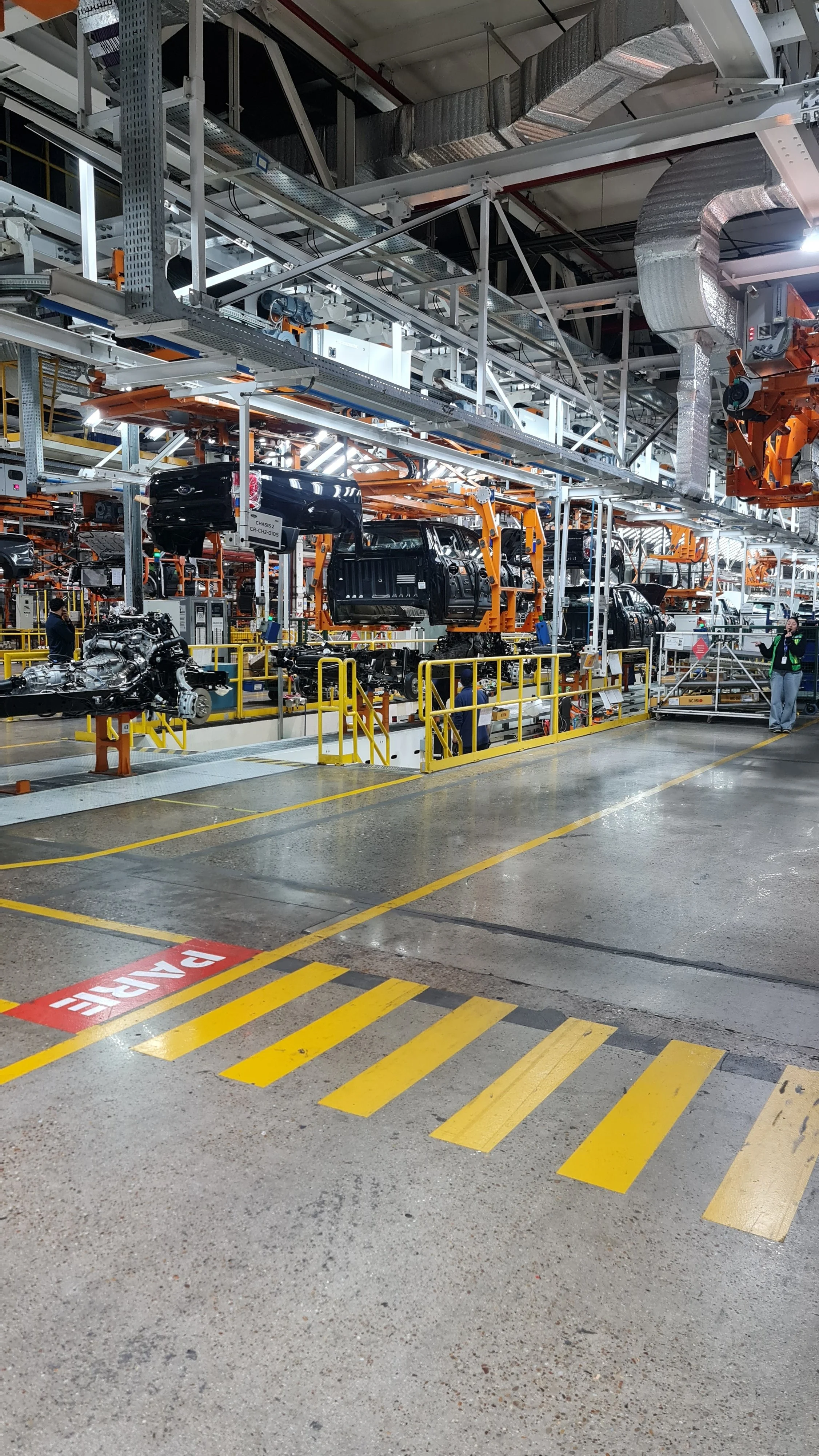 Fábrica de ensamblaje automotriz con vehículos en proceso de fabricación, trabajadores y maquinaria en un entorno industrial.