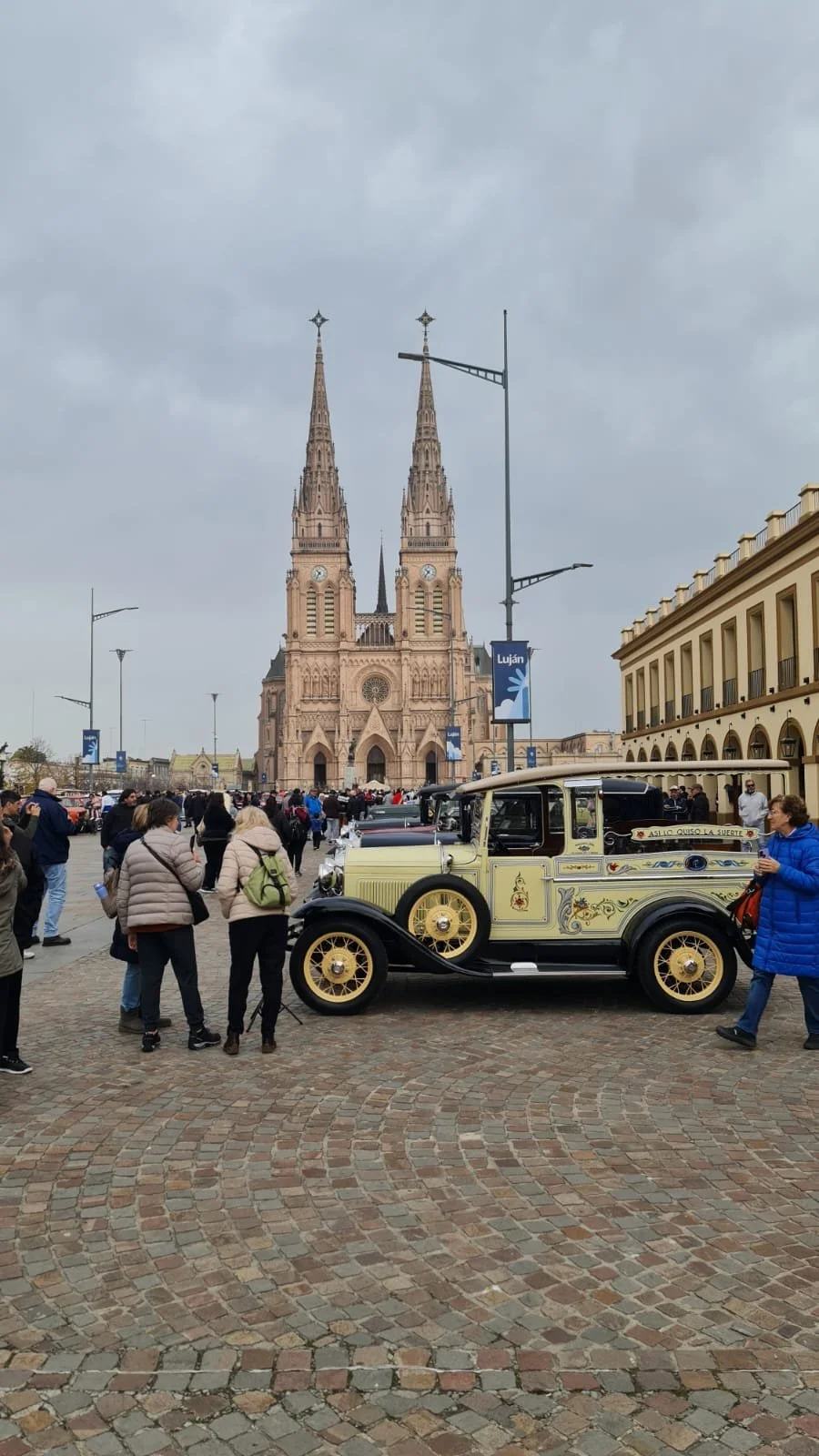 Vista de una plaza con una iglesia gótica de fondo y un coche vintage en primer plano, rodeado de personas.