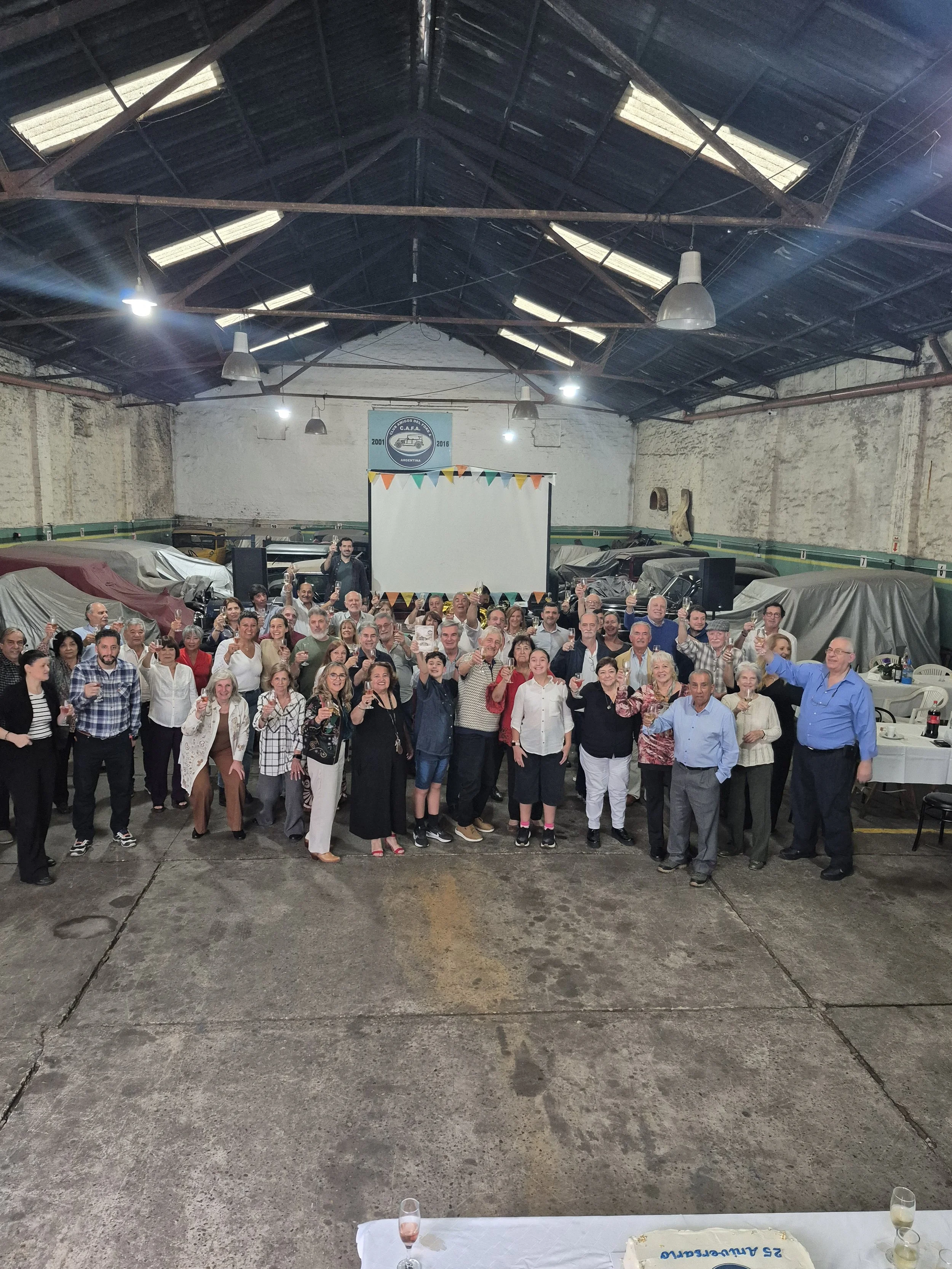 Grupo de personas celebrando en un salón amplio con paredes de ladrillo y techo de lámina, en una fiesta con una mesa con pastel y bebidas al frente.