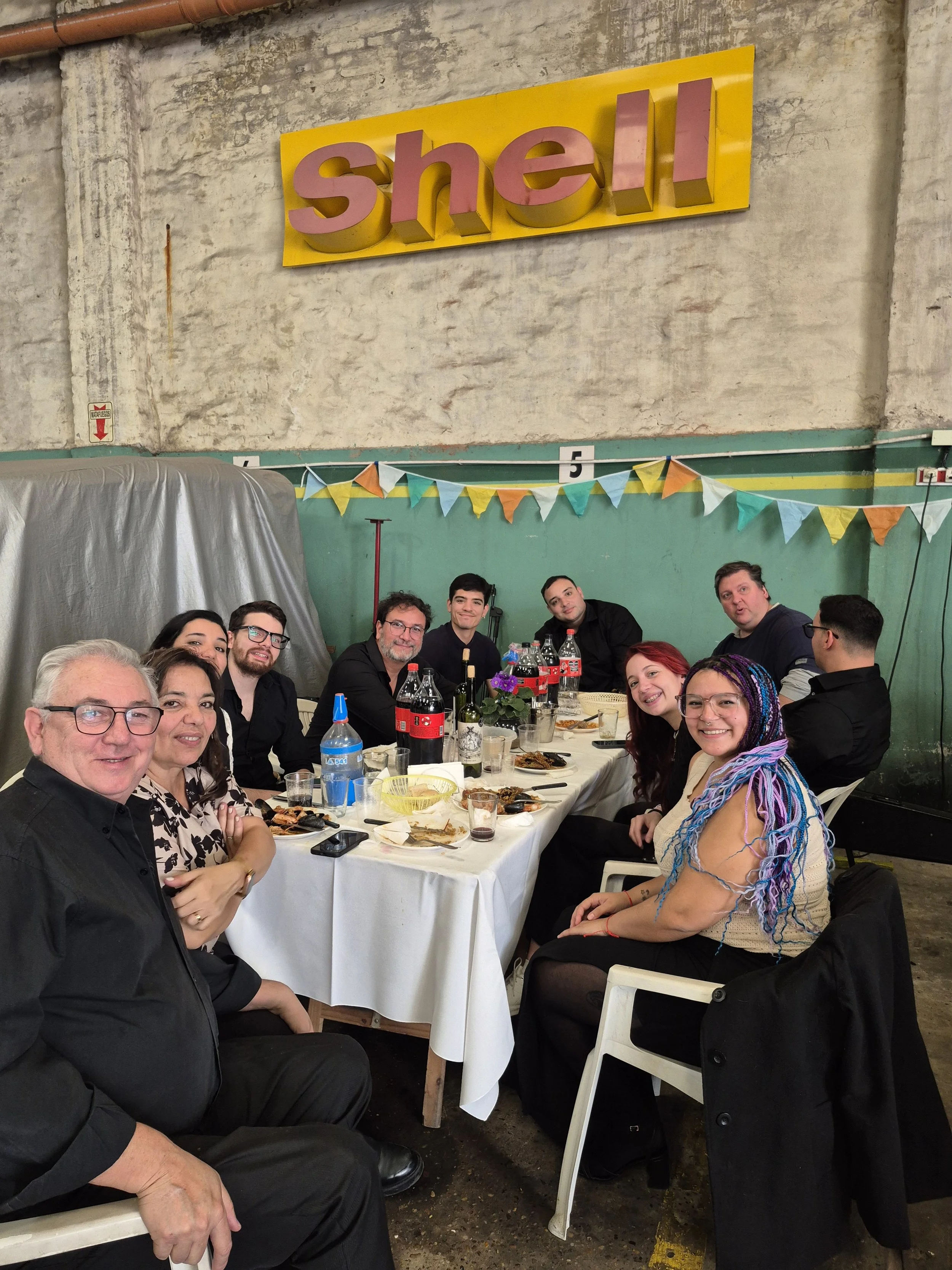 Grupo de personas en una comida o celebración, sentadas alrededor de una mesa con comida y refrescos en un espacio con paredes de concreto y decoraciones de banderines de colores.