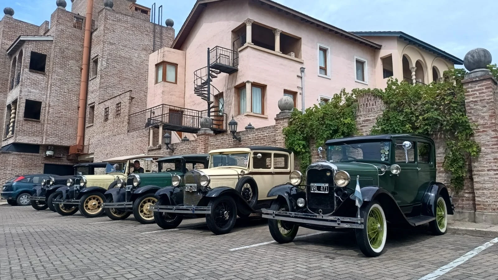 Cinco autos clásicos estacionados en una calle de adoquines, con un edificio de fondo que tiene paredes de ladrillo, balcones y una escalera de espiral en el exterior.
