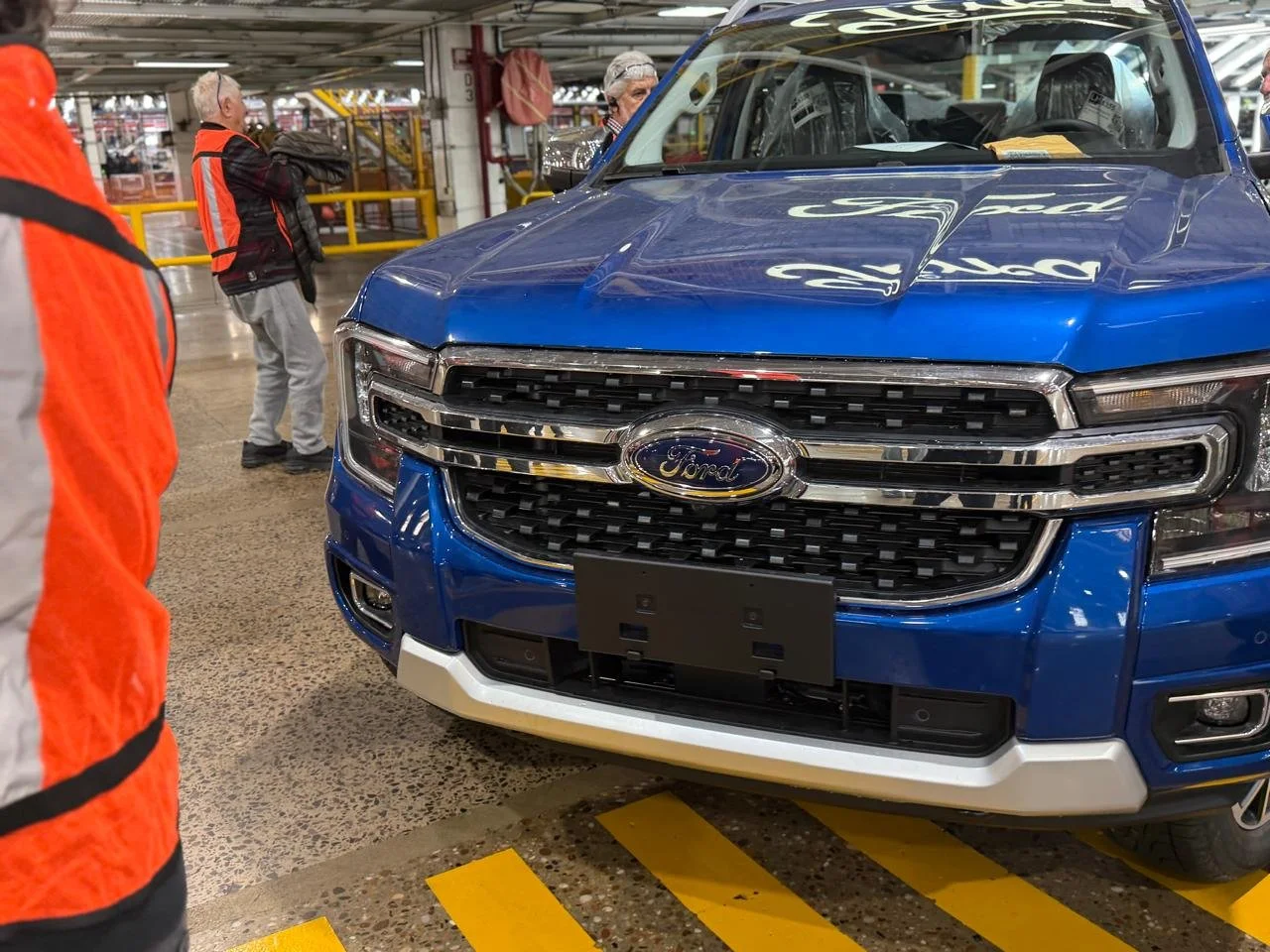 Vista frontal de un automóvil Ford azul en una fábrica o almacén, con varias personas observando y enmarcado por estructuras metálicas y señalización en el piso.