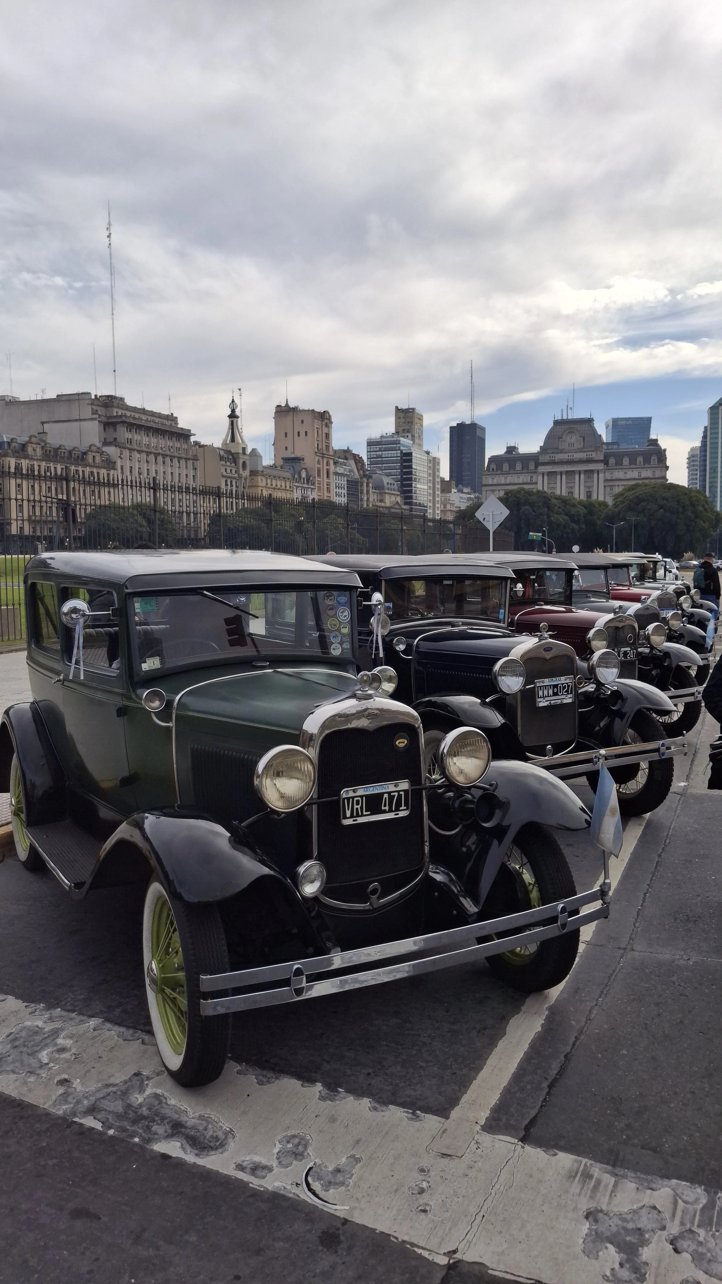 Autos antiguos en exhibición en una ciudad, con edificios altos y nubes en el cielo de fondo.