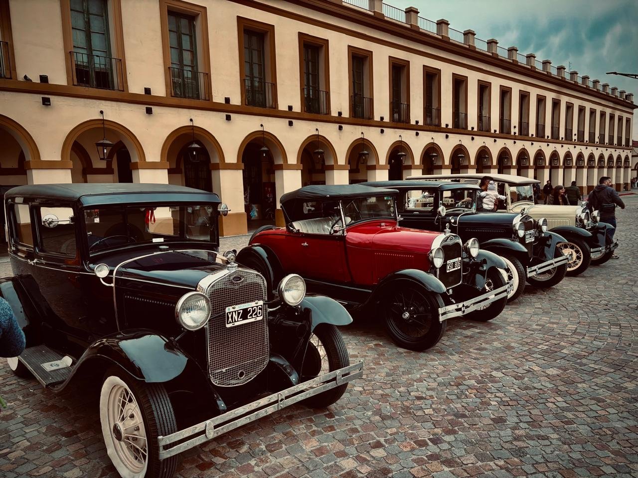 Carros clásicos en una calle adoquinada frente a un edificio de colores claros con arcos y ventanas grandes.