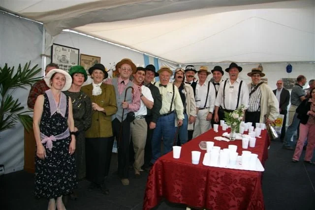 Grupo de personas vestidas con ropa vintage en una fiesta o evento bajo una carpa, con una mesa cubierta con un mantel rojo y adornos.