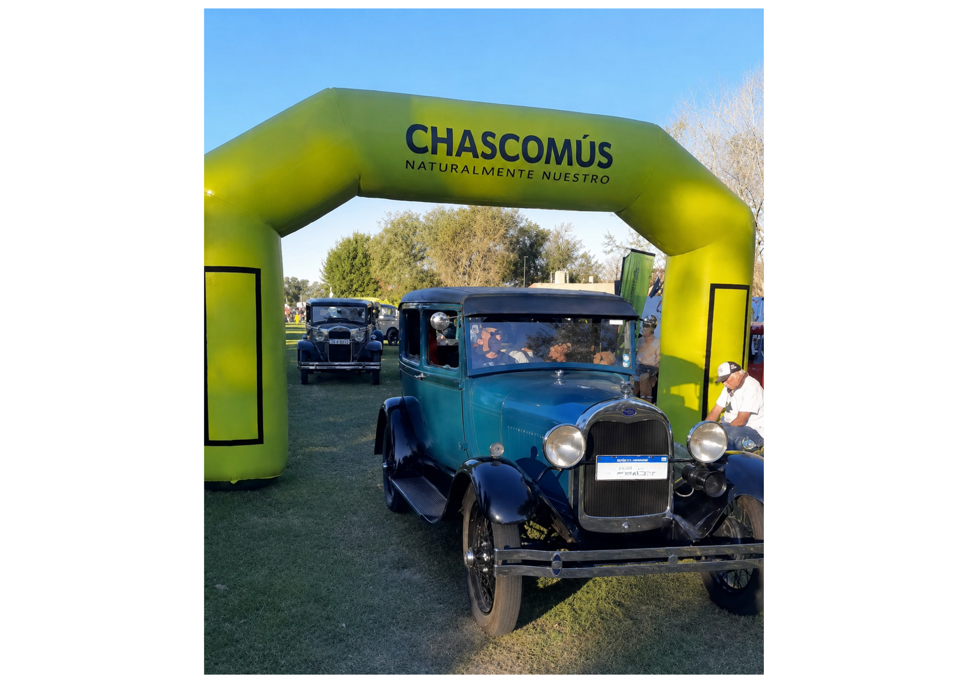 Autos antiguos en un evento, con un arco inflable verde que tiene la palabra "Chascomús" y el lema "Naturalmente Nuestro" en letras negras, en un día soleado con árboles y cielo despejado.