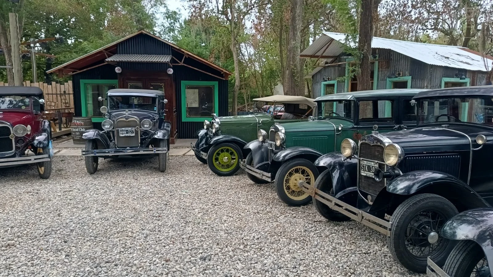Varios autos clásicos antiguos estacionados en un terreno con grava, con casas de madera al fondo y árboles alrededor.