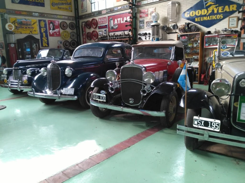 Tres autos clásicos en un taller o museo, rodeados de herramientas y anuncios de neumáticos como Goodyear y otros, con paredes llenas de mapas y repisas con piezas y accesorios automotrices.
