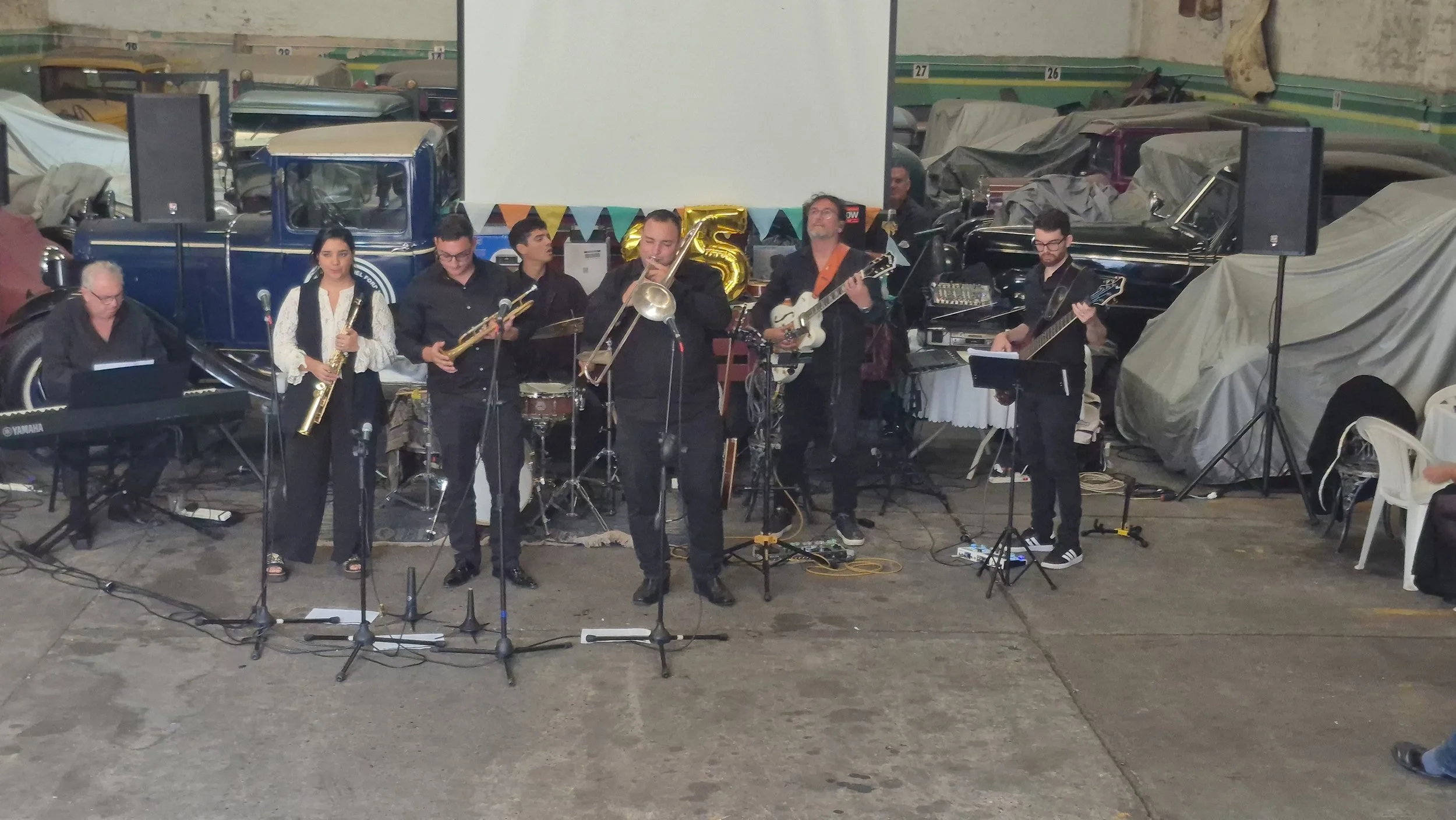 A un grupo de músicos tocando instrumentos en un escenario, con fondo decorado con banderines y globos dorados, y varios autos cubiertos con sábanas.