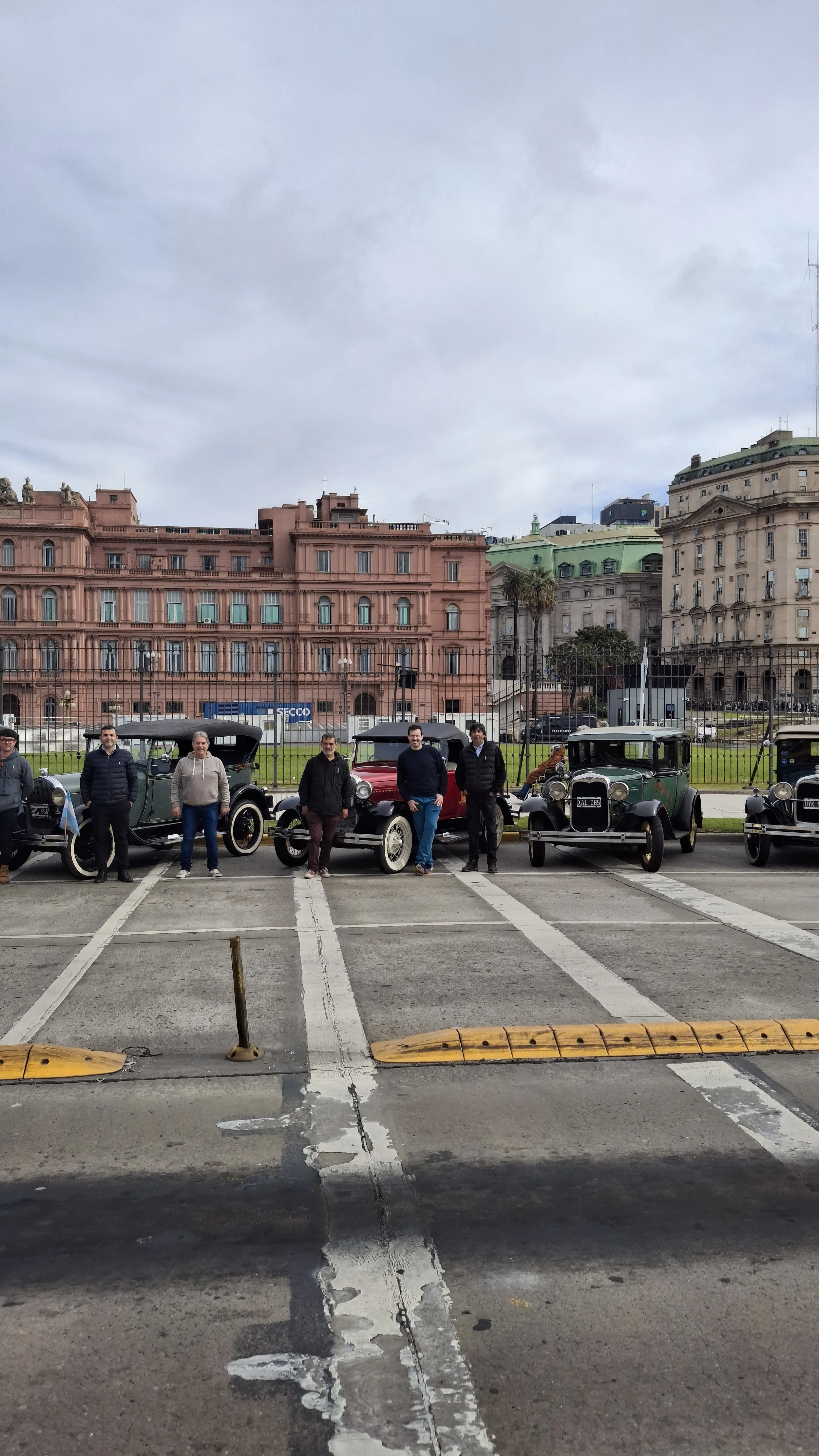 Cinco personas de pie junto a autos antiguos en un parqueadero, con edificios históricos al fondo en un día nublado.