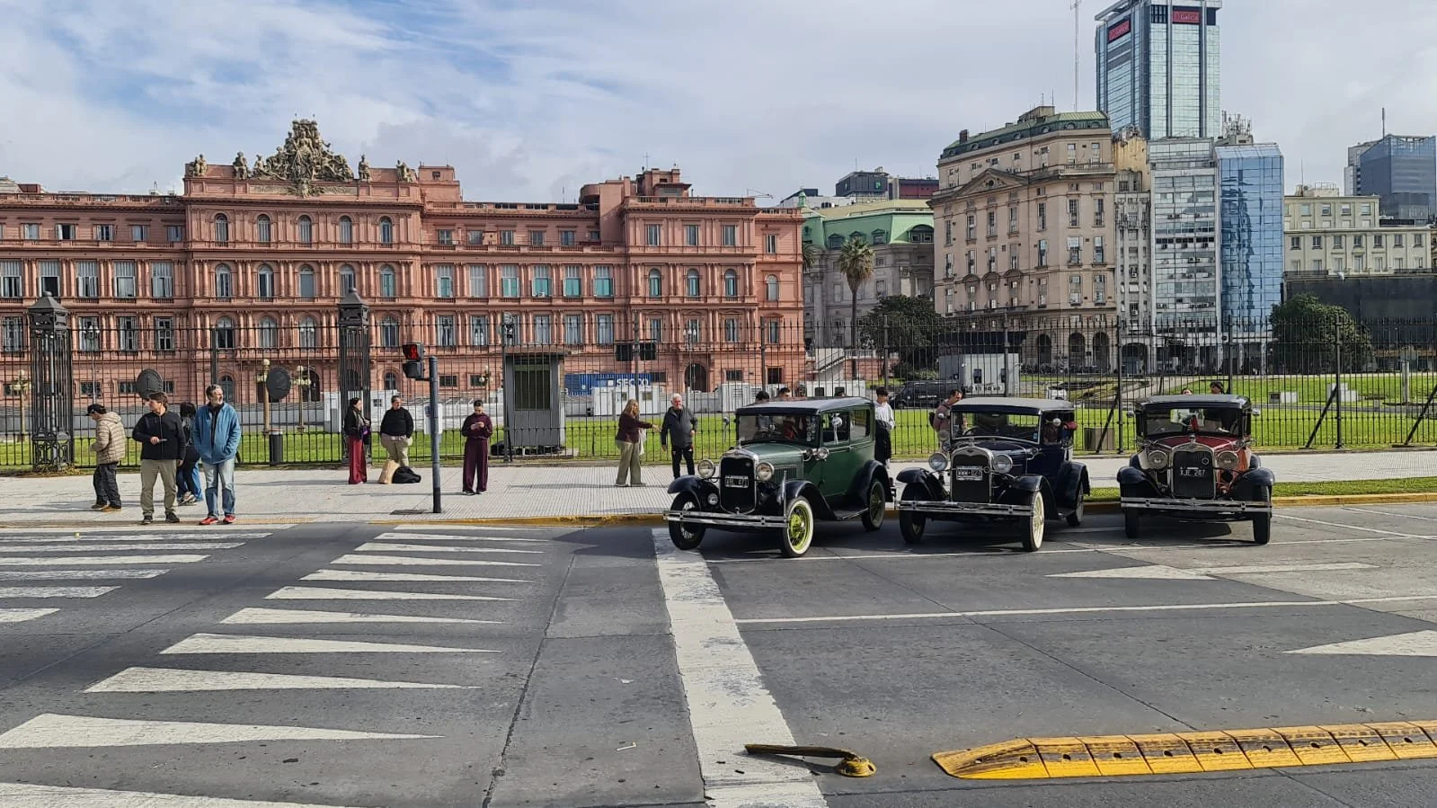 Cinco autos clásicos de color negro y verde estacionados en la calle frente a un parque con reja y edificios antiguos y modernos en el fondo, con personas caminando y cruzando la calle.