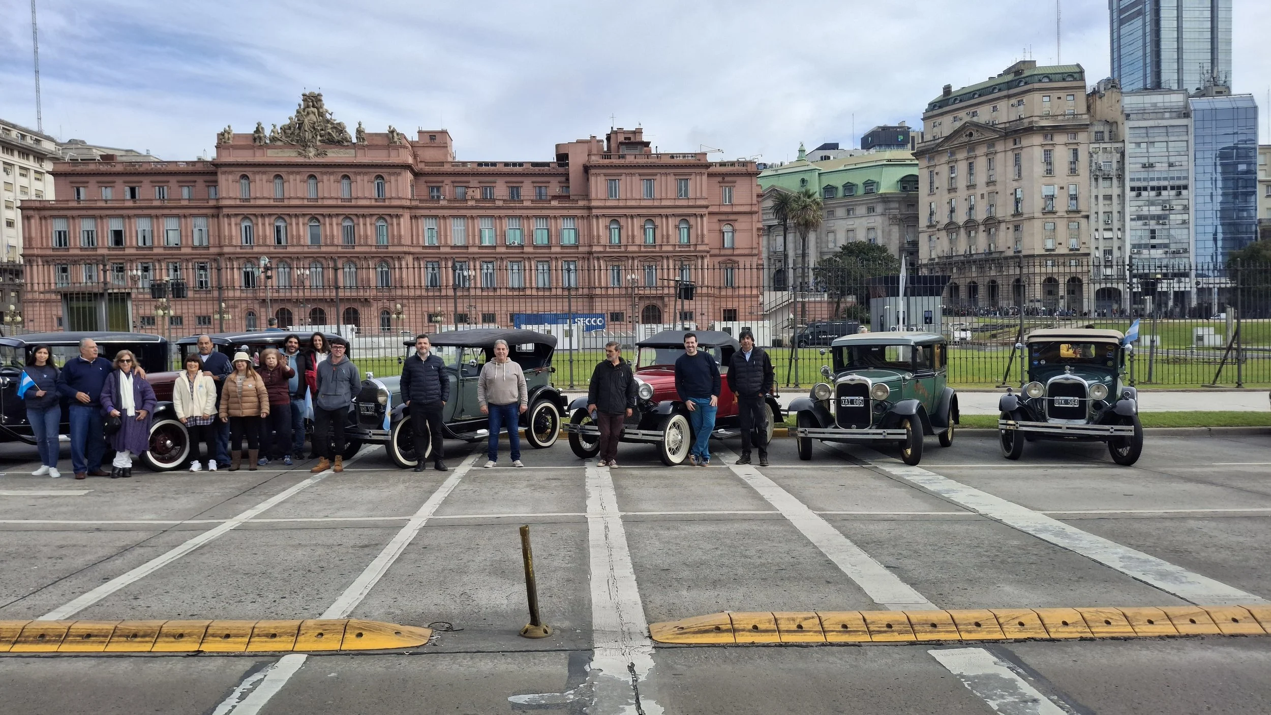 Grupo de personas junto a autos antiguos en una calle, con edificios históricos y modernos en el fondo.