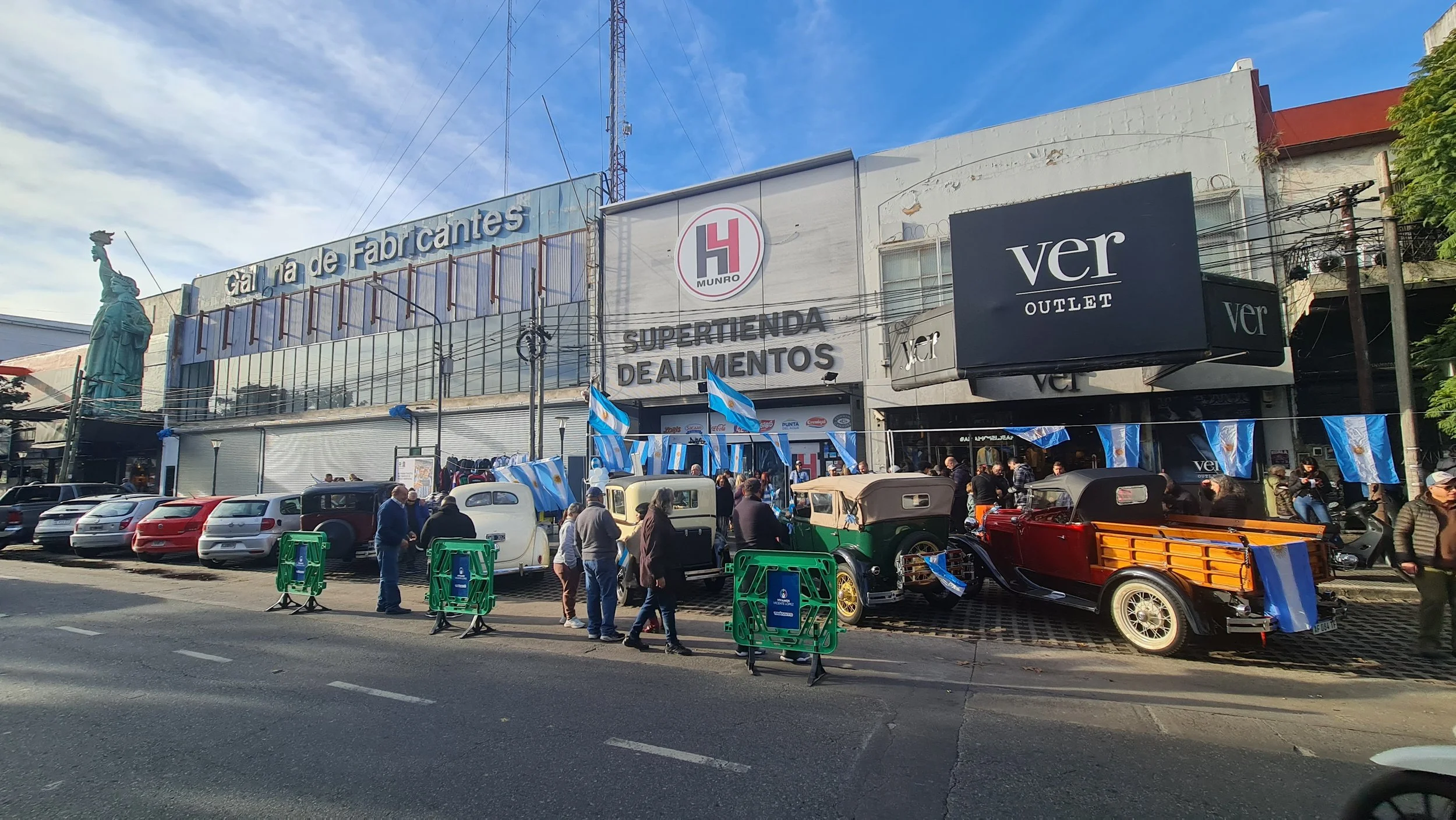 Feria o evento en la calle con coches antiguos, banderas argentinas, y un grupo de personas en la acera frente a un edificio con señalización que dice 'Galería de Fabricantes' y 'Supertienda de Alimentos'.