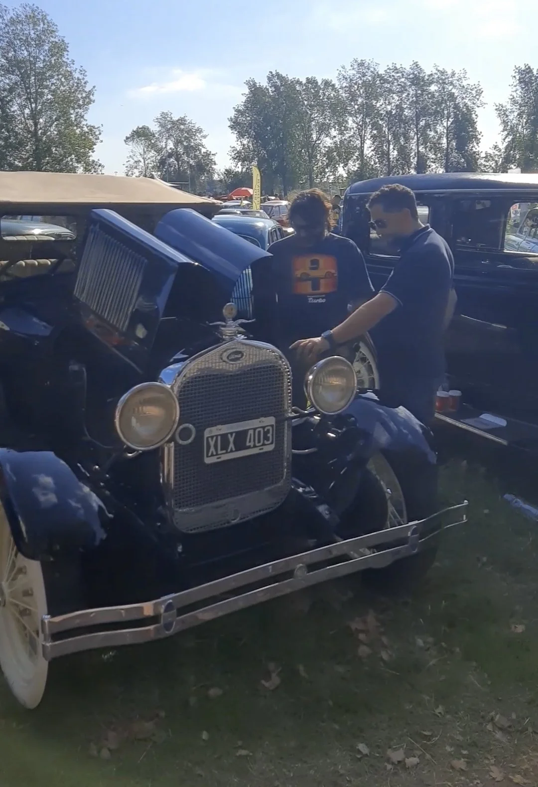 Un auto antiguo de color negro, con matrícula XLX 403, exhibido en un evento de autos clásicos, con dos personas observando la máquina.