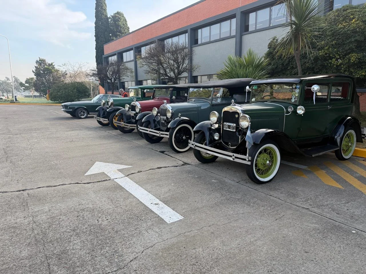 Cinco autos clásicos estacionados en línea en un aparcamiento, con un edificio y árboles de fondo.