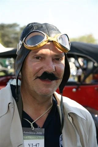 Hombre disfrazado con gafas de aviador, bigote falso y gorra de piloto, en un evento o reunión al aire libre.