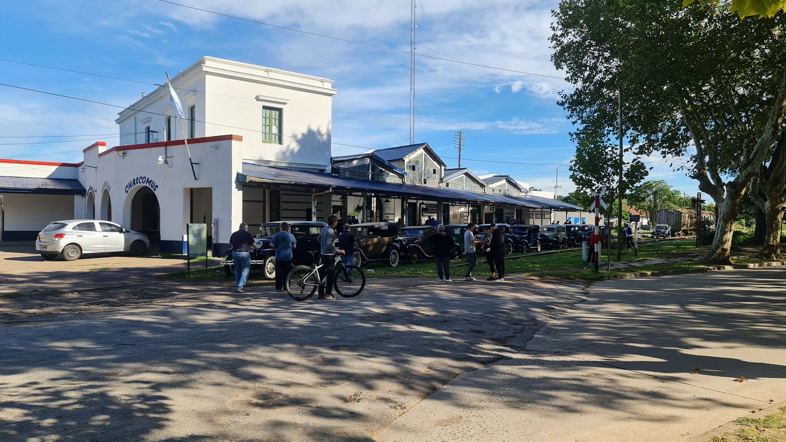 Vista del museo Chascomús con varias personas y autos vintage frente a la entrada en un día soleado con cielo despejado y árboles grandes en el parque cercano.