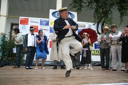 Grupo de personas vestidos con ropa vintage participando en una danza o acto cultural en un escenario al aire libre, con un hombre en el centro bailando con sombrero.