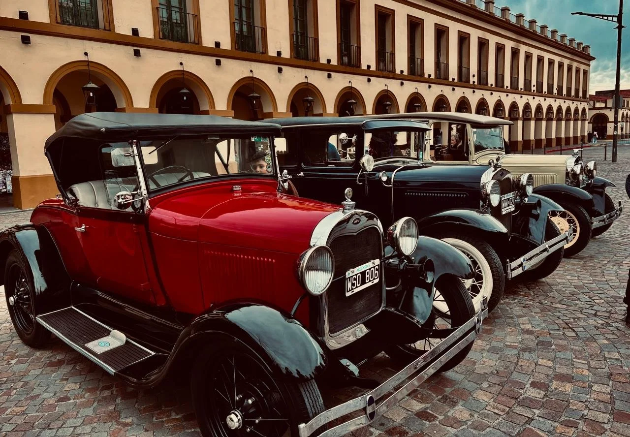 Tres autos antiguos estacionados en una plaza pavimentada, con un edificio de arcos en el fondo.