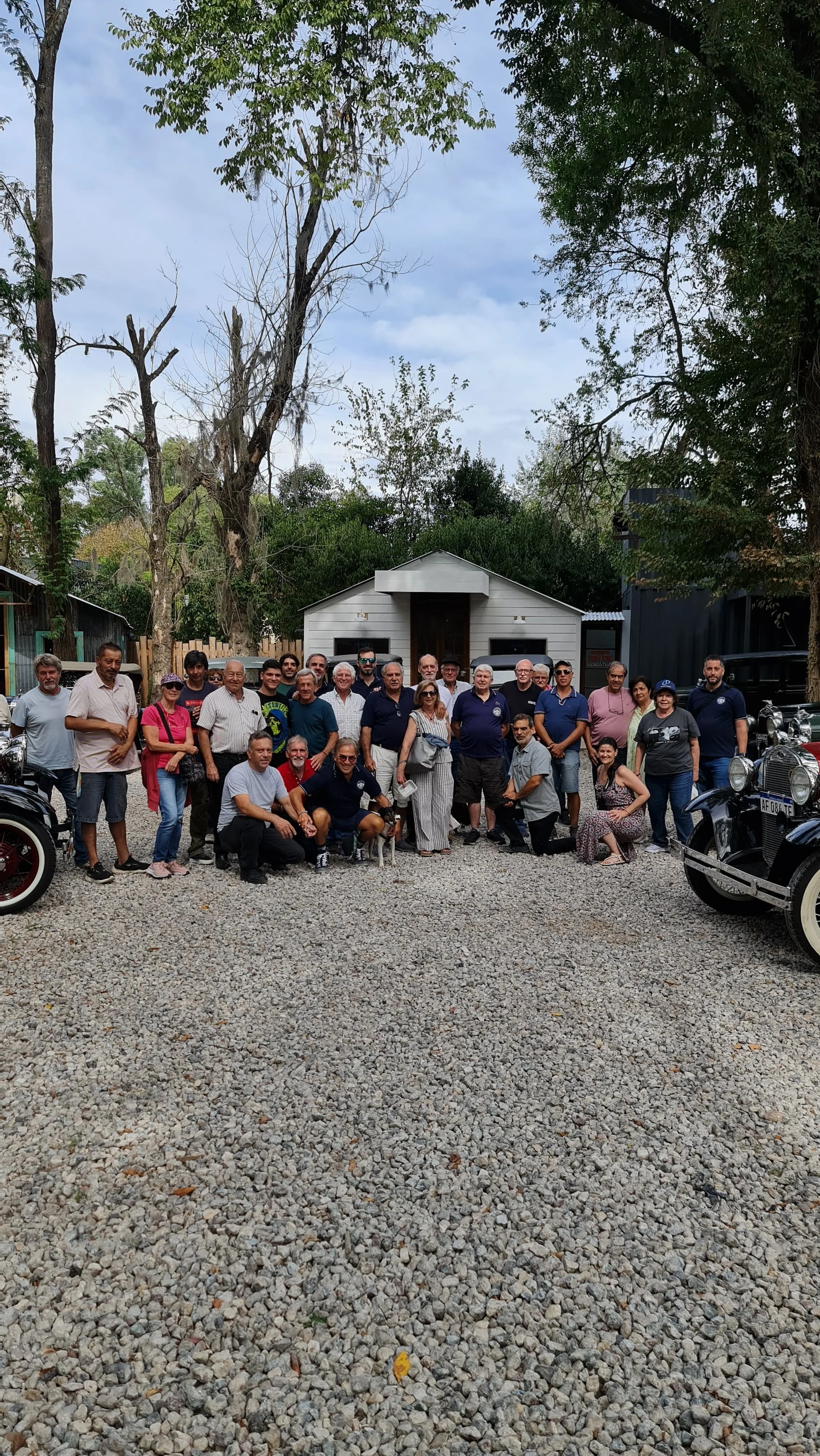 Grupo de personas reunidas frente a una casa blanca de madera, con árboles altos y autos antiguos en el entorno.