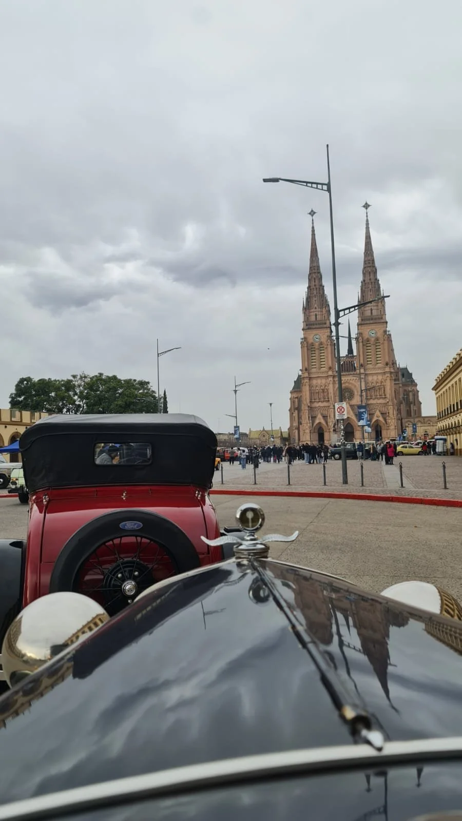 Vista desde adelante de coches antiguos en una plaza frente a una iglesia gótica con torres altas y un reloj, en un día nublado.