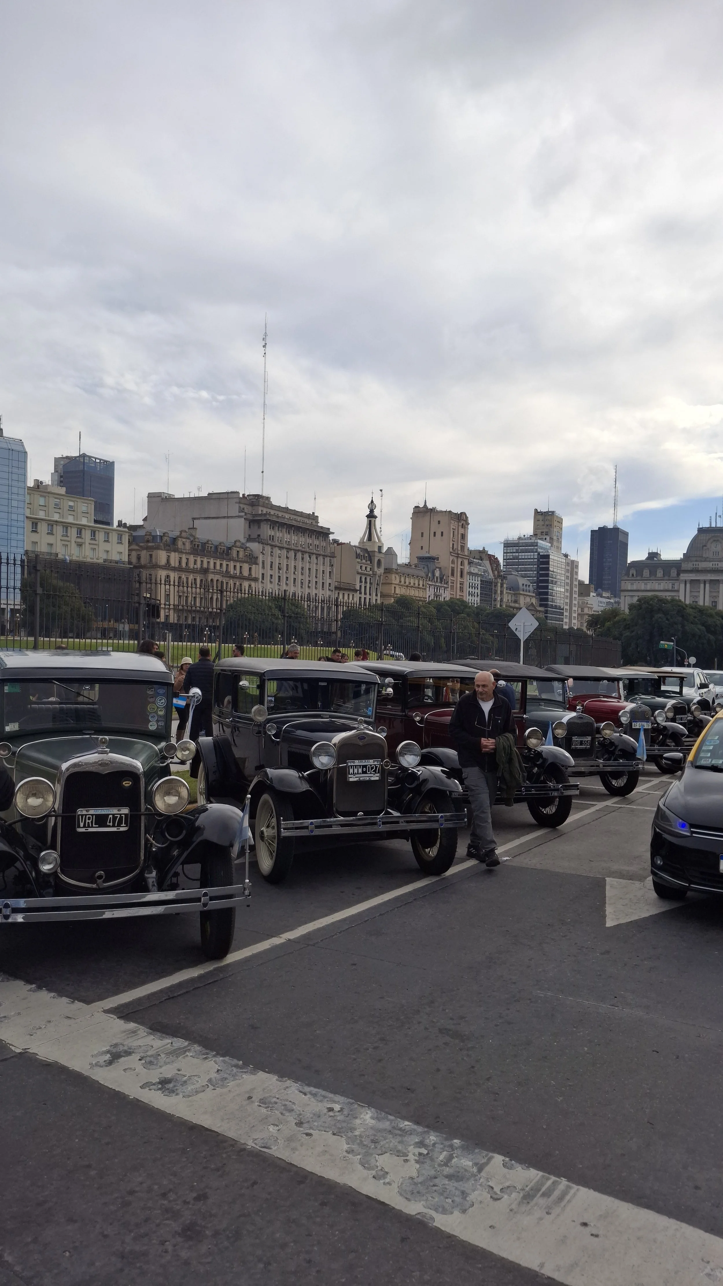Aparcamiento con autos antiguos en una ciudad grande, con edificios en el fondo y un cielo nublado.