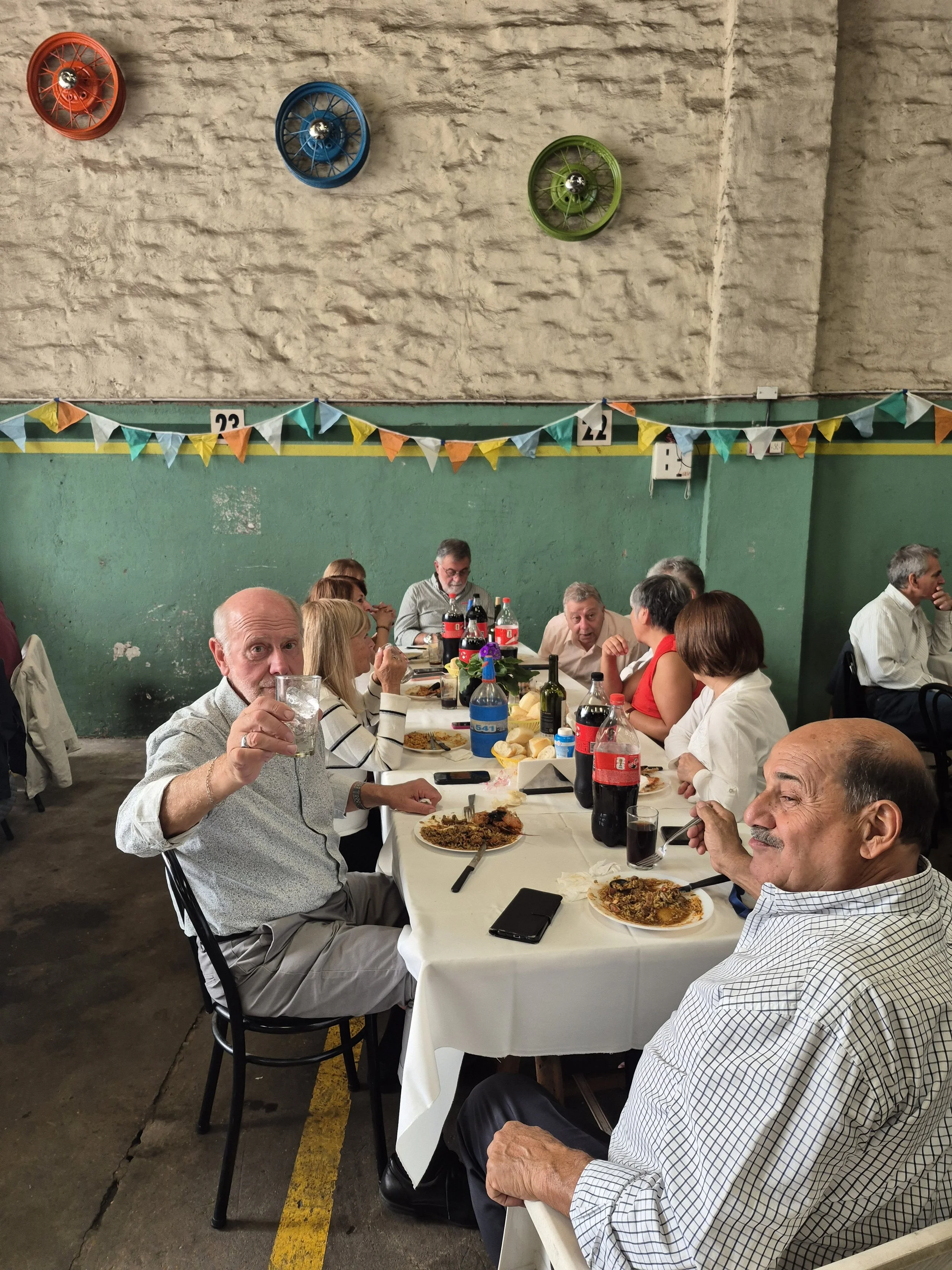 Personas mayores comiendo en una reunión social en un salón con paredes verdes y decoraciones de banderines de colores, con comida y bebidas en la mesa.