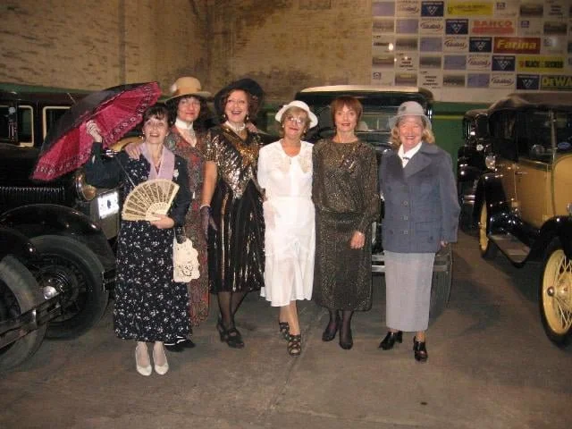Grupo de seis mujeres vestidas con ropa vintage, posando frente a autos antiguos en un estacionamiento.