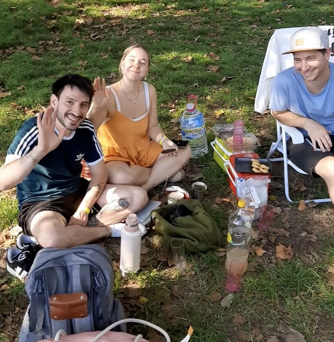 Tres personas disfrutando un picnic en un parque, dos sentados en el suelo y una en una silla de playa, con comida y botellas en el área.