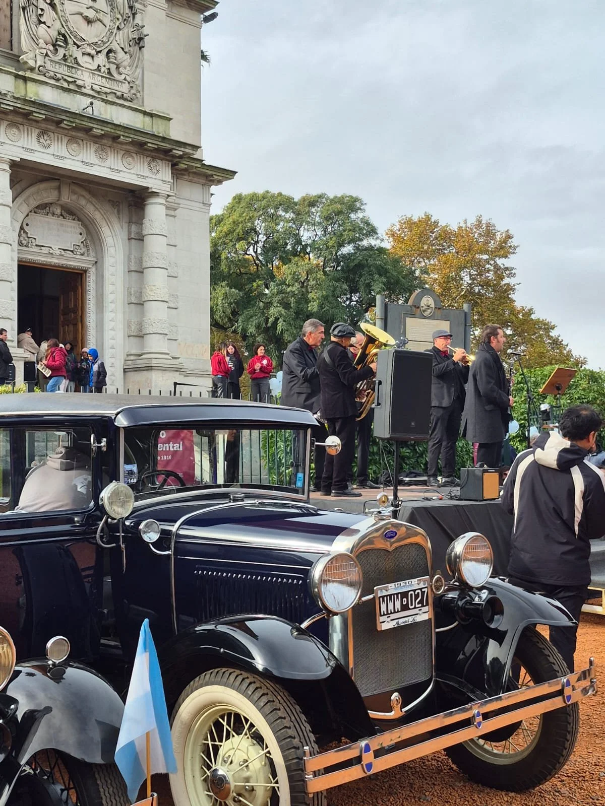 Un automóvil clásico negro en primer plano y un grupo de músicos tocando en un escenario al fondo, frente a un edificio emblemático, en un día nublado con árboles en el fondo.