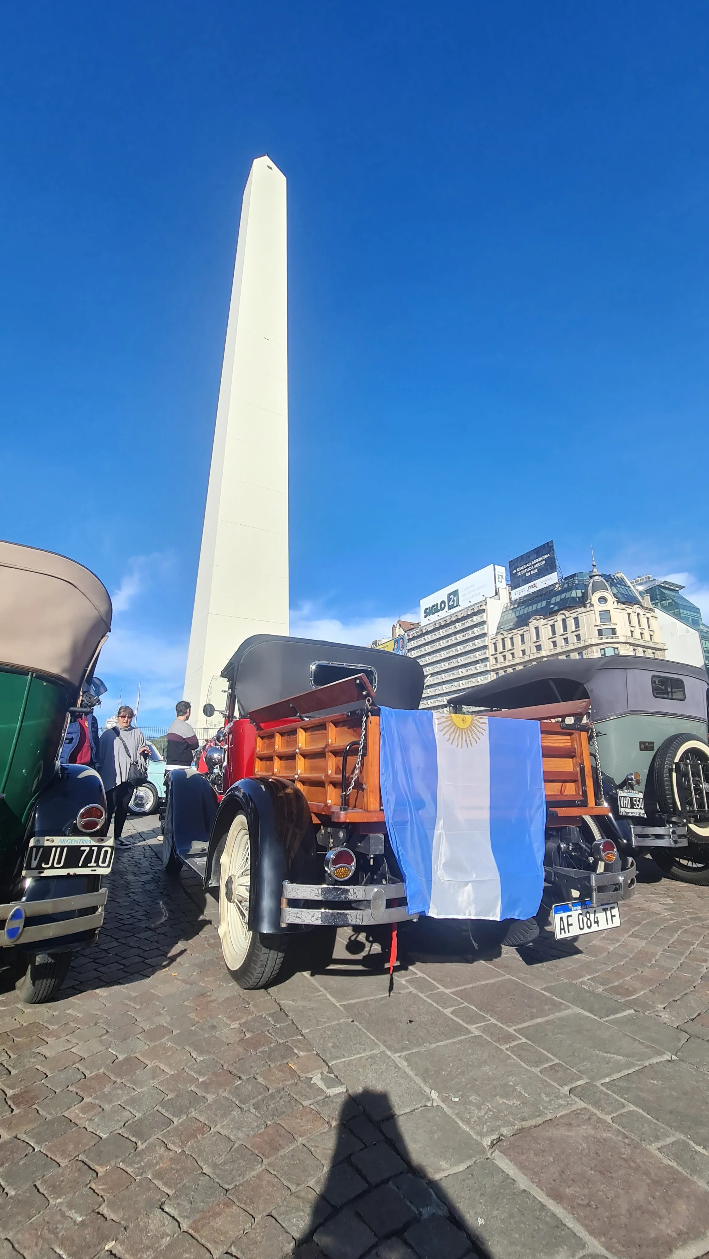 Vehículos antiguos en una plaza, uno con una bandera de Argentina. En el fondo, un obelisco y edificios urbanos. Personas caminando y en motocicleta.