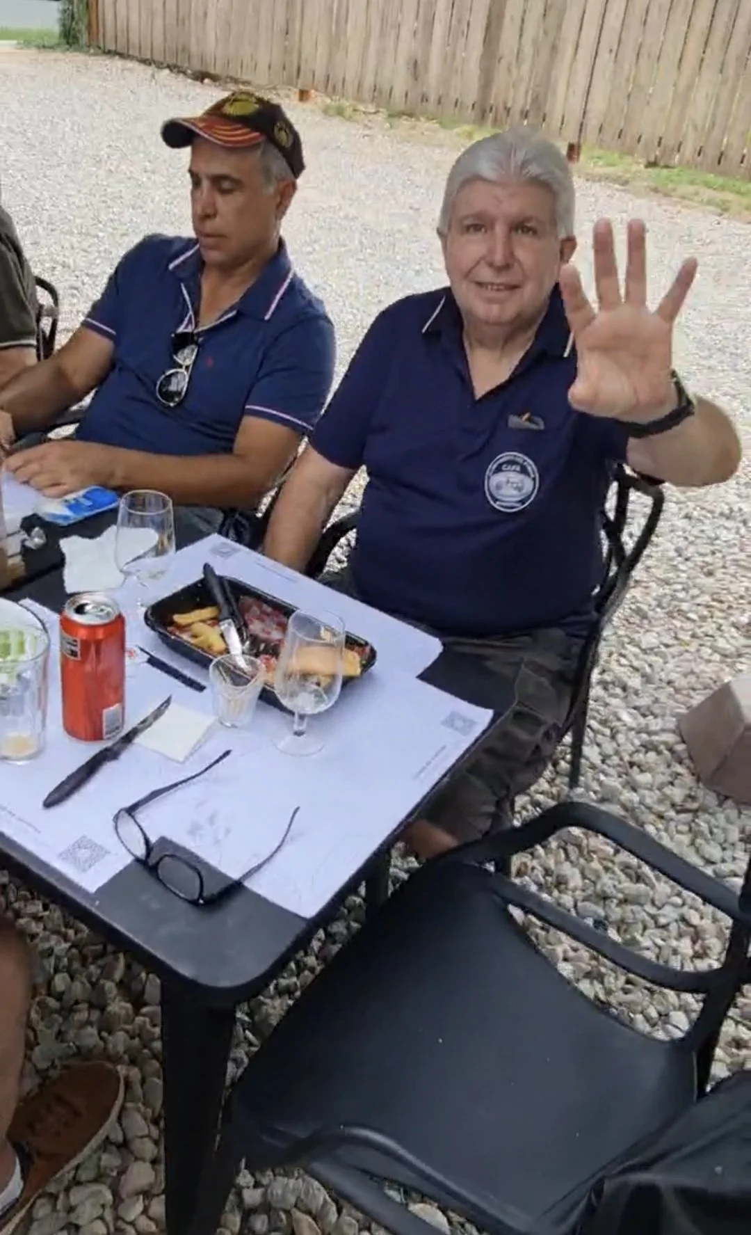 Hombre con cabello gris y camisa azul sentados en una mesa de reunión al aire libre, haciendo una señal de saludo con la mano derecha, acompañado de otra persona de cabello oscuro y camisa de color azul oscuro, en un entorno exterior con valla de mad