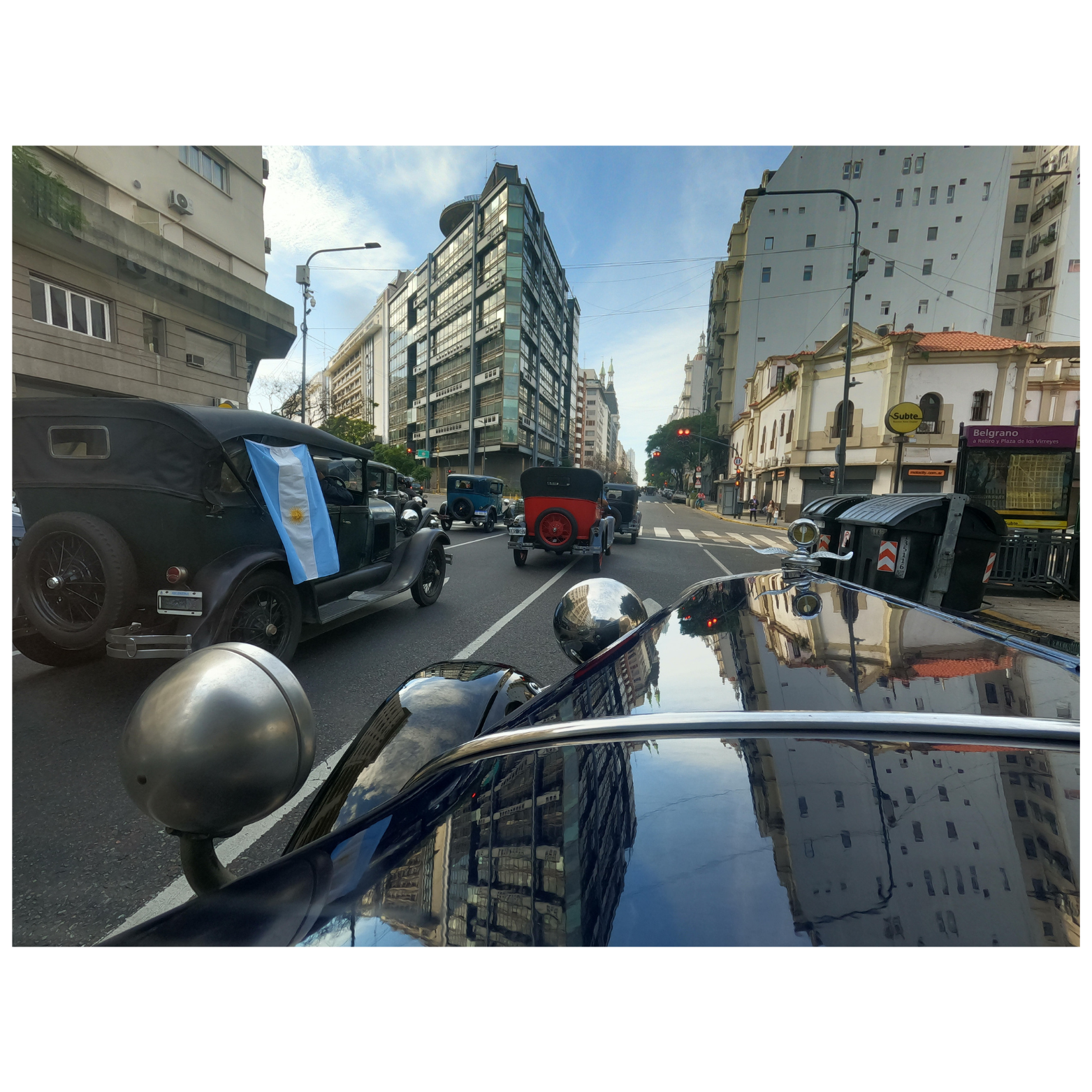 Vista de una calle con autos antiguos, incluyendo uno con bandera argentina, y edificios altos en el fondo, en una ciudad de Buenos Aires.