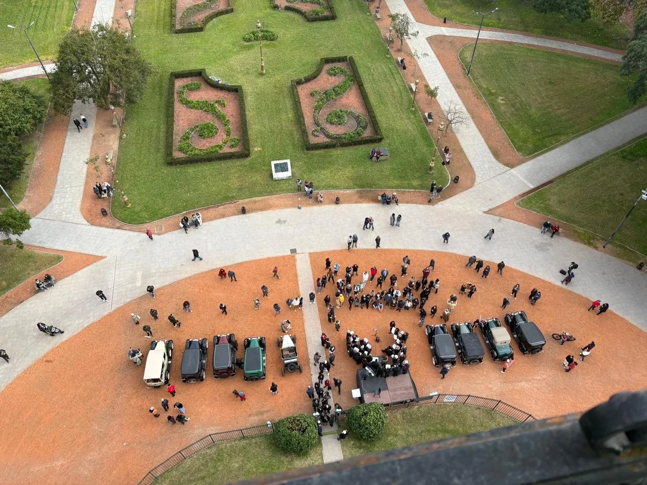 Vista aérea de una plaza con mucha gente, carros estacionados y áreas verdes con jardines y caminos pavimentados.