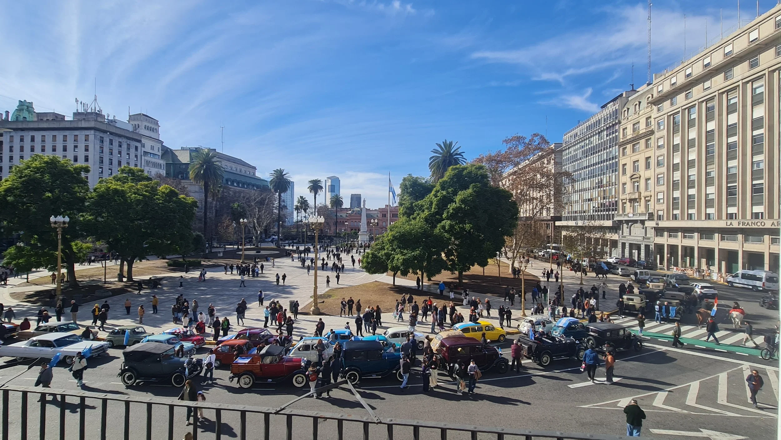 Plaza concurrida en una ciudad con edificios altos y palmeras, con muchas personas caminando y carros estacionados y en movimiento.