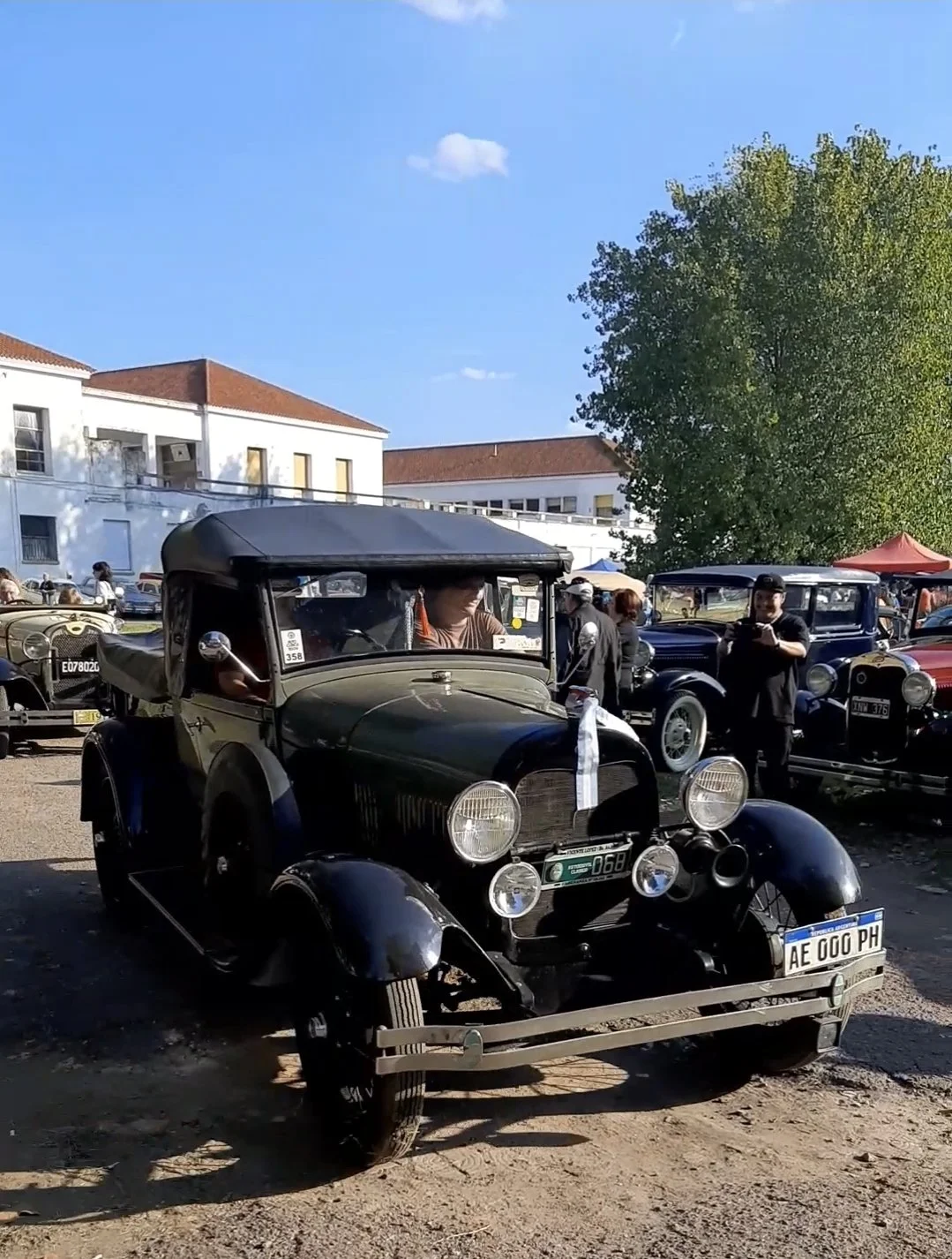 Autos clásicos en una exhibición al aire libre, con edificios blancos y árboles verdes de fondo, y varias personas observando y tomando fotos.