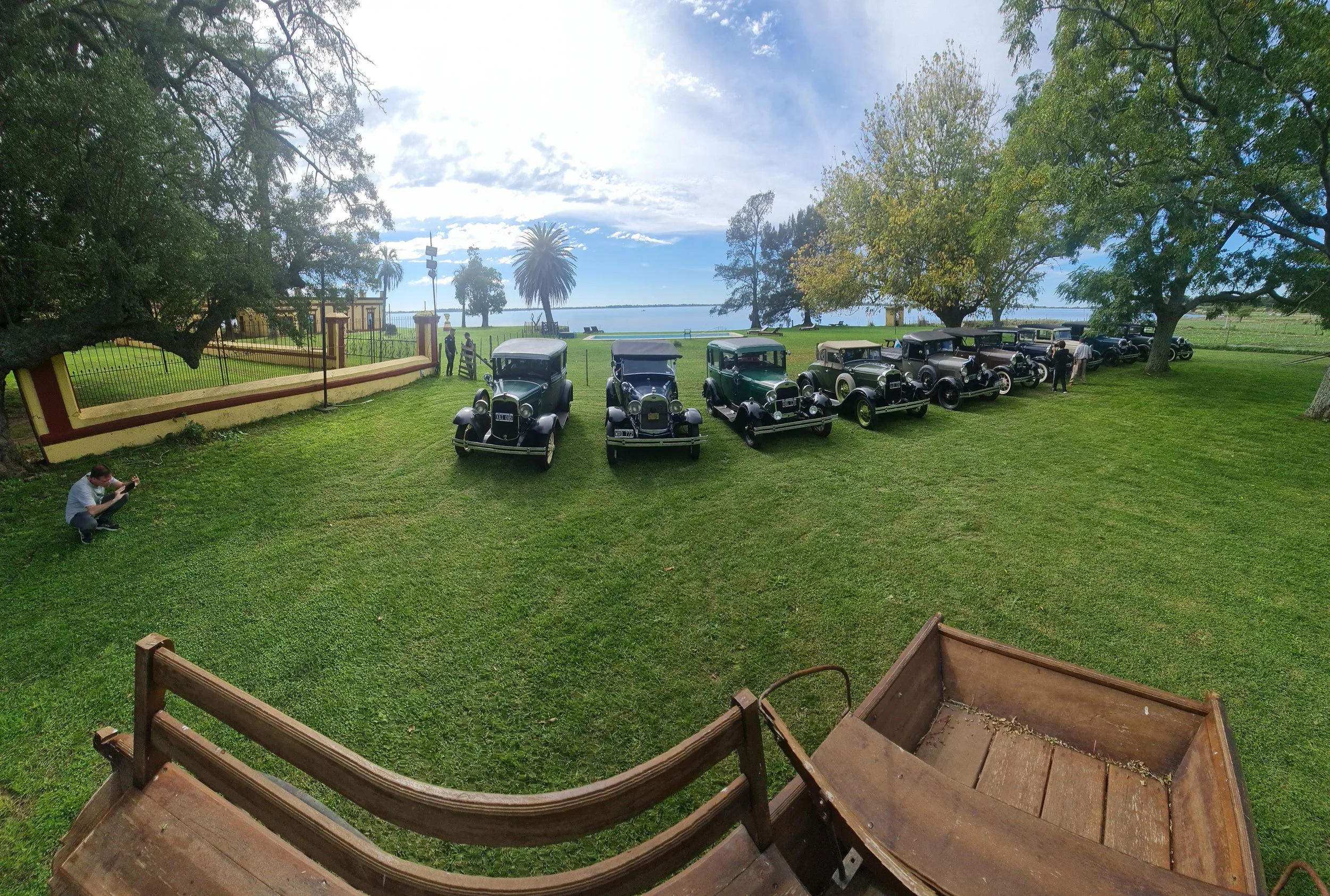 Vista de coches clásicos estacionados en un parque con árboles y vista al lago, bajo un cielo parcialmente nublado.