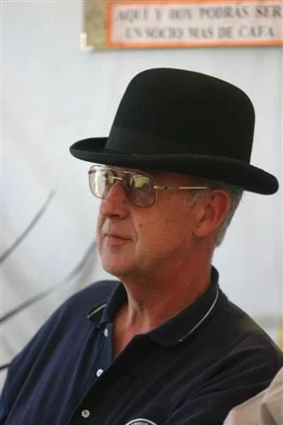 Hombre con sombrero negro y gafas, vestido con camiseta polo azul, en un entorno interior.
