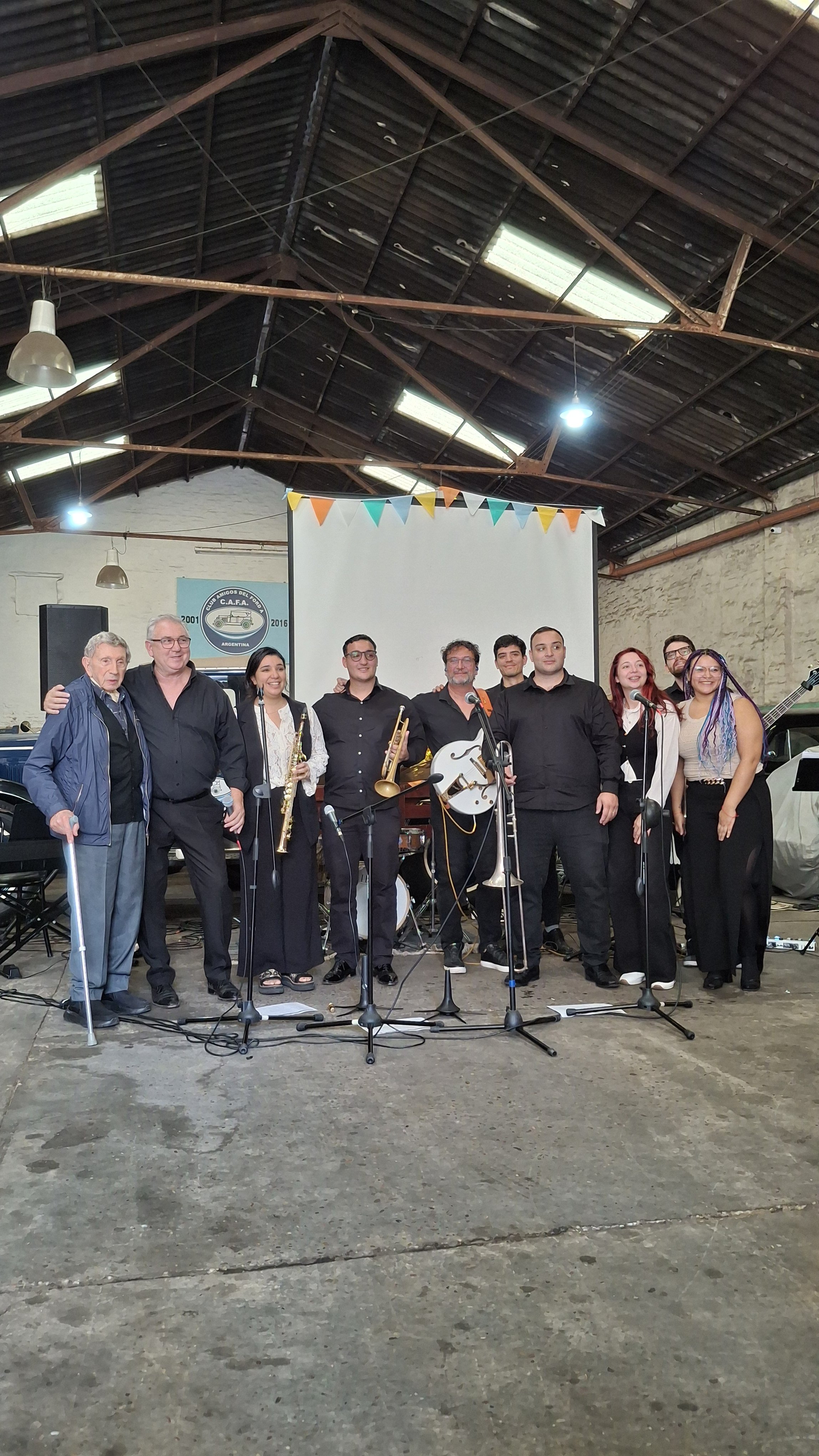 Grupo de personas en una sala con techos altos, algunas con instrumentos musicales, posando para una foto. Fondo con un cartel y una pantalla. Decoraciones de banderines en la parte superior.
