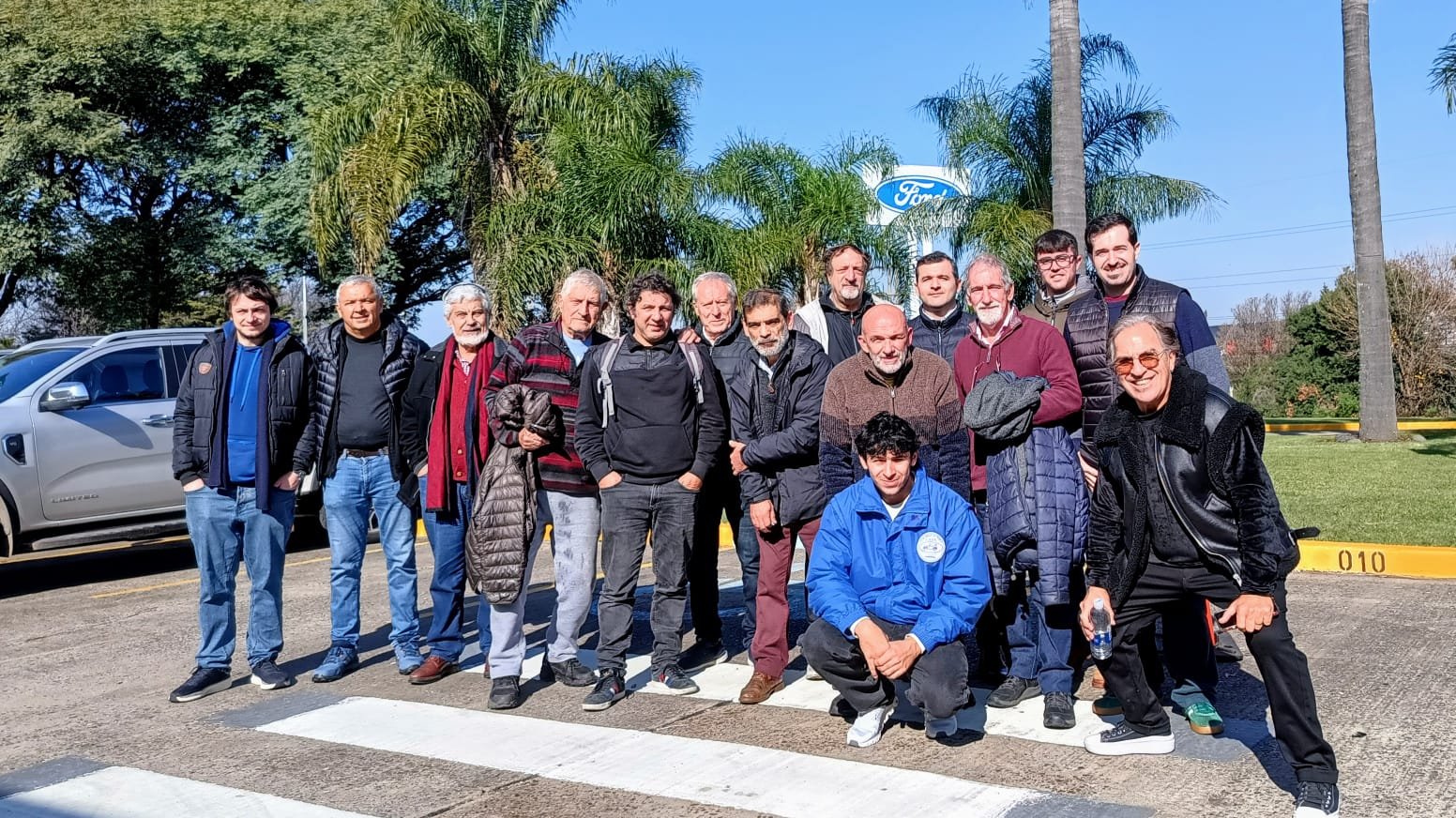 Grupo de personas posando para una foto al aire libre sobre un paso peatonal, con árboles y un cartel de Ford en el fondo.