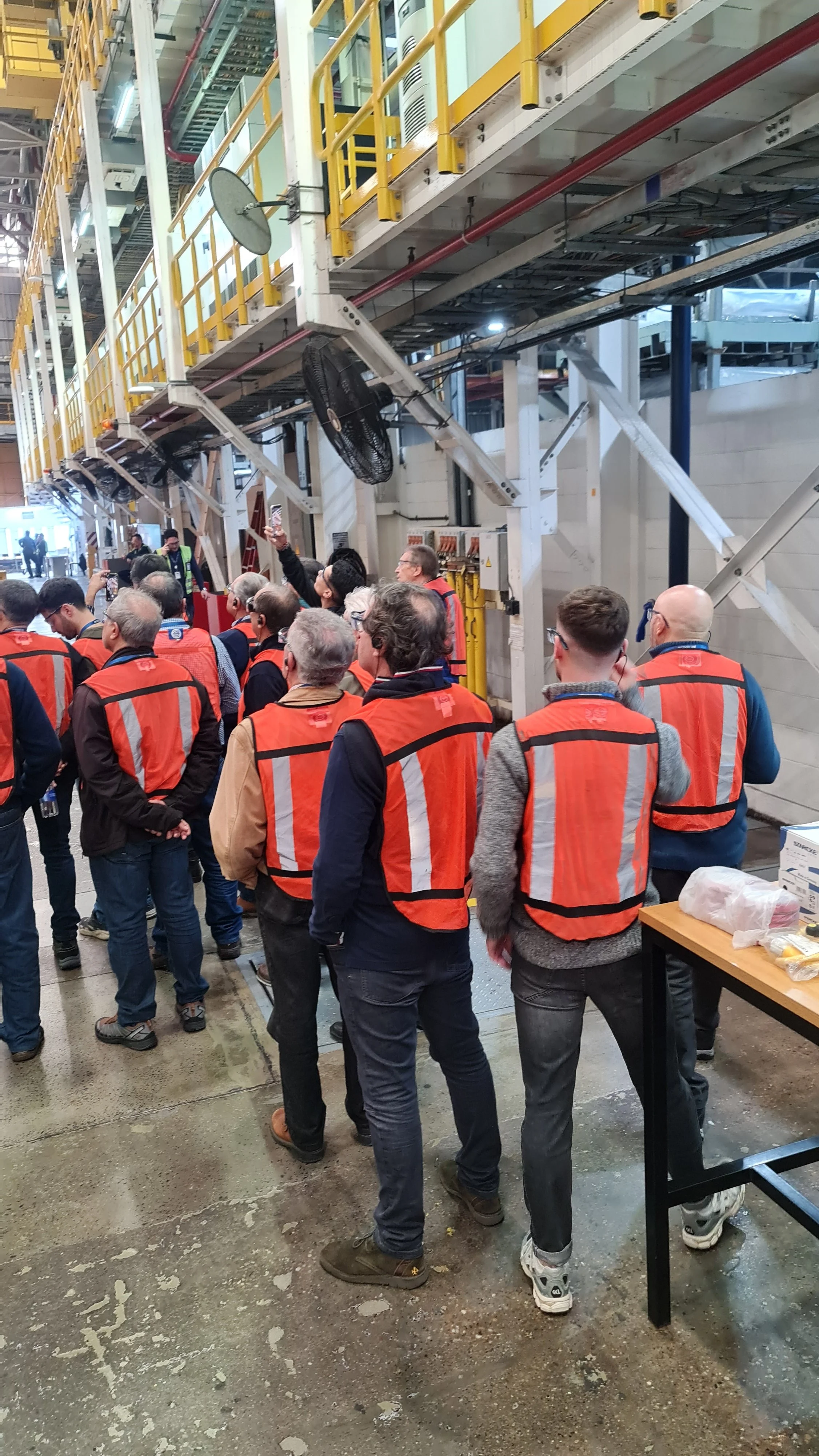 Grupo de personas con chalecos de seguridad naranjas en una planta industrial, observando y tomando fotos de las instalaciones.