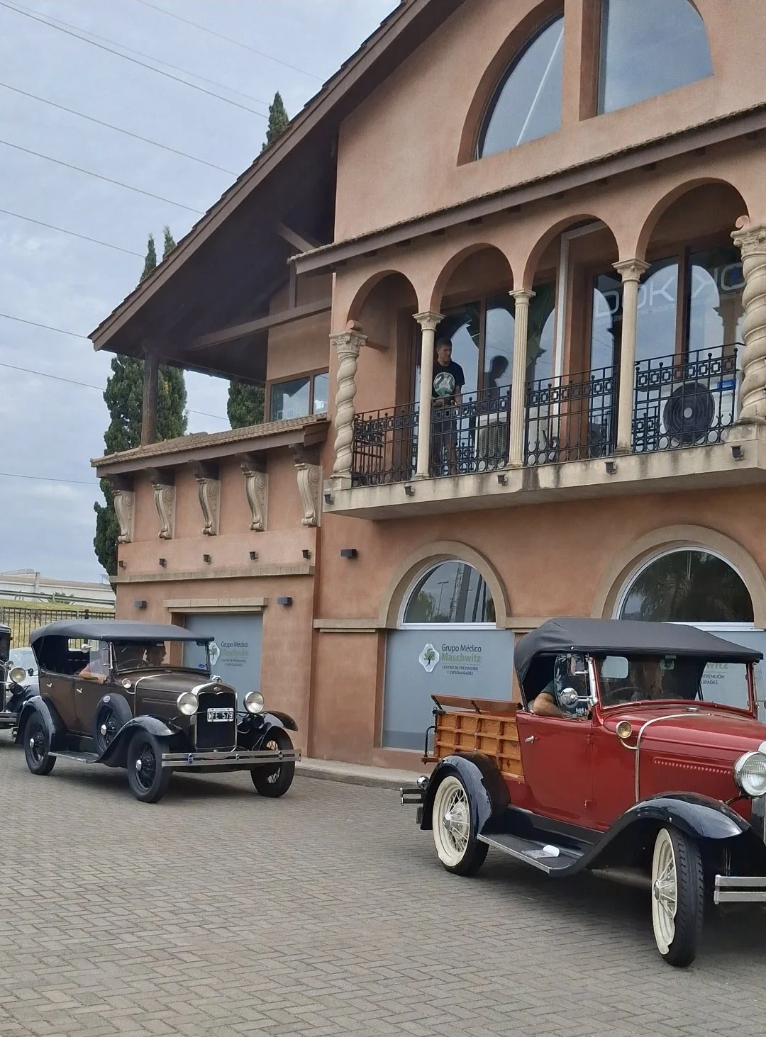 Dos autos antiguos estacionados frente a un edificio de color terracota con ventanas y balcones con columnas, y una persona observando desde el balcón.