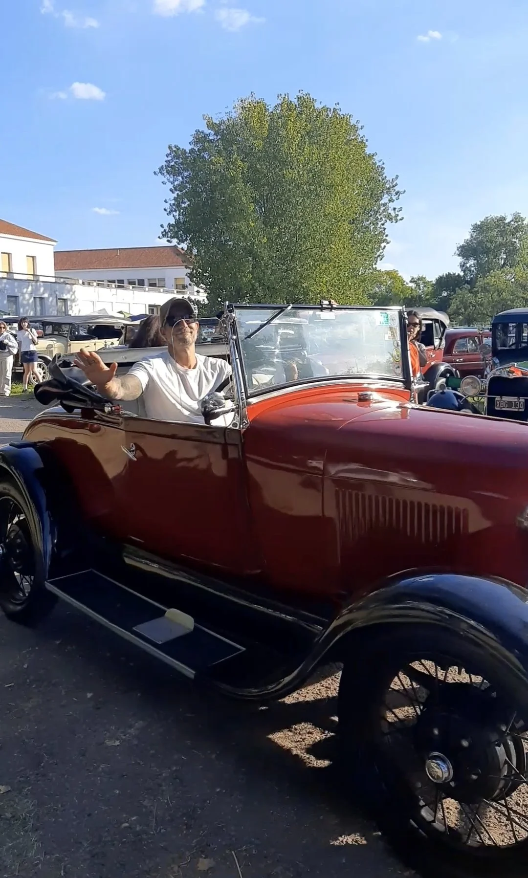 Hombre conduciendo un coche clásico de color rojo en un evento al aire libre, sonriendo y saludando con la mano derecha mientras lleva gafas de sol y gorra, en un día soleado con árboles y otras personas y coches vintage en el fondo.