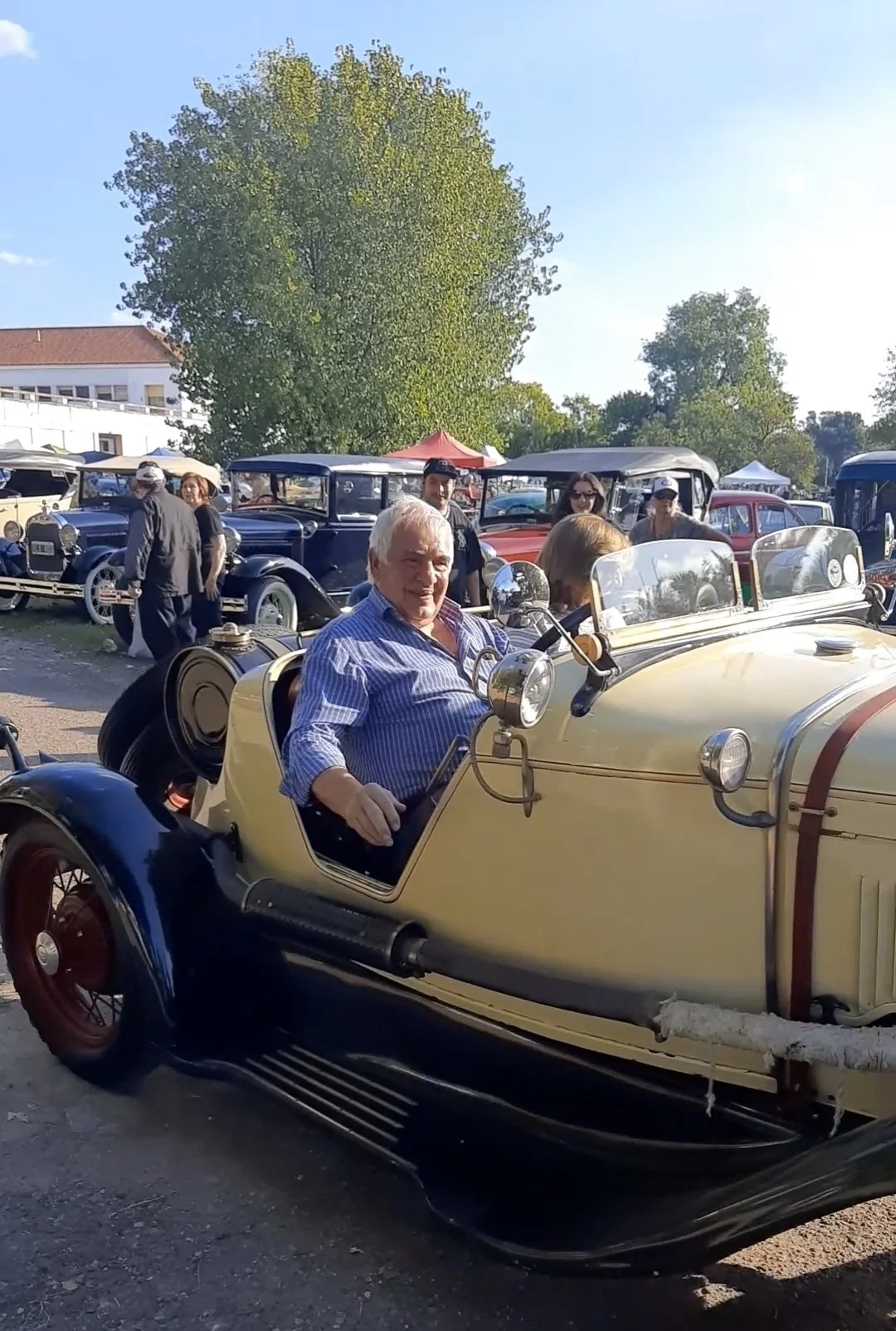 Hombre sentado en coche antiguo de color amarillo en exhibición con varias personas y otros autos clásicos en el fondo en un día soleado.