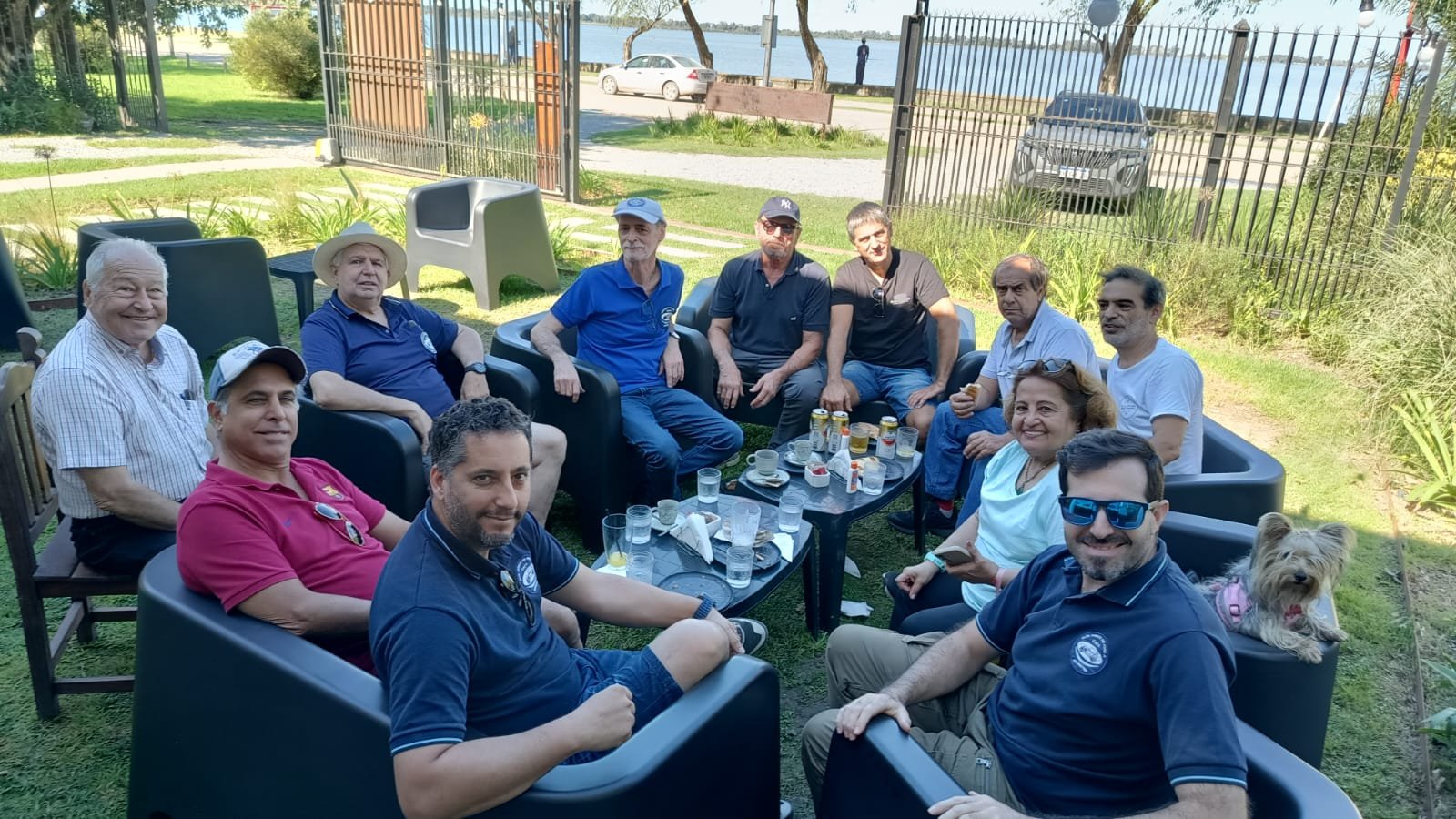 Grupo de personas reunidas en un jardín disfrutando de bebidas y risas, con vista al lago al fondo.