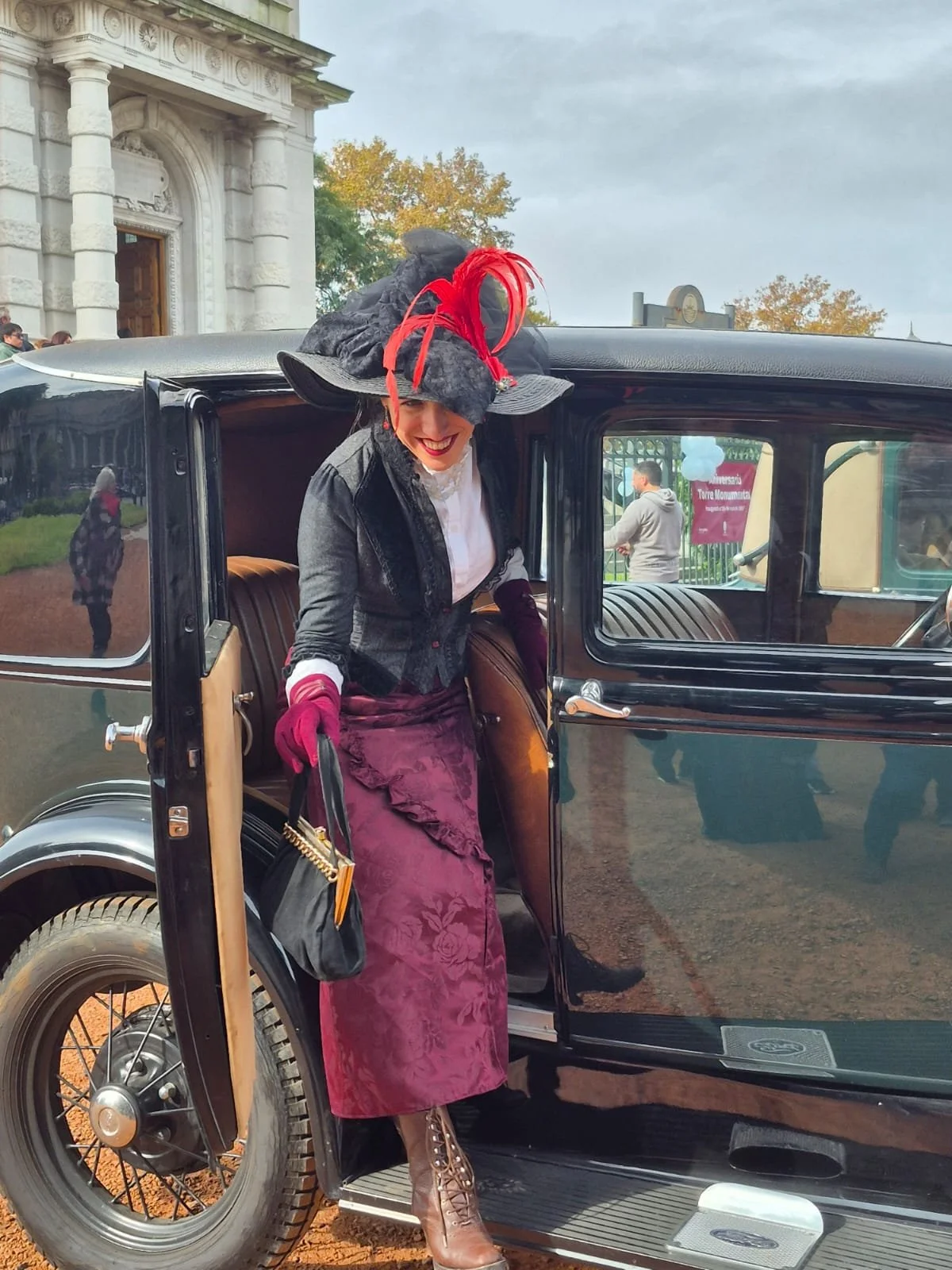 Una mujer con vestimenta vintage y sombrero elegante sale de un automóvil antiguo negro, en un entorno que parece ser una visita turística a un monumento histórico en un día nublado o lluvioso.