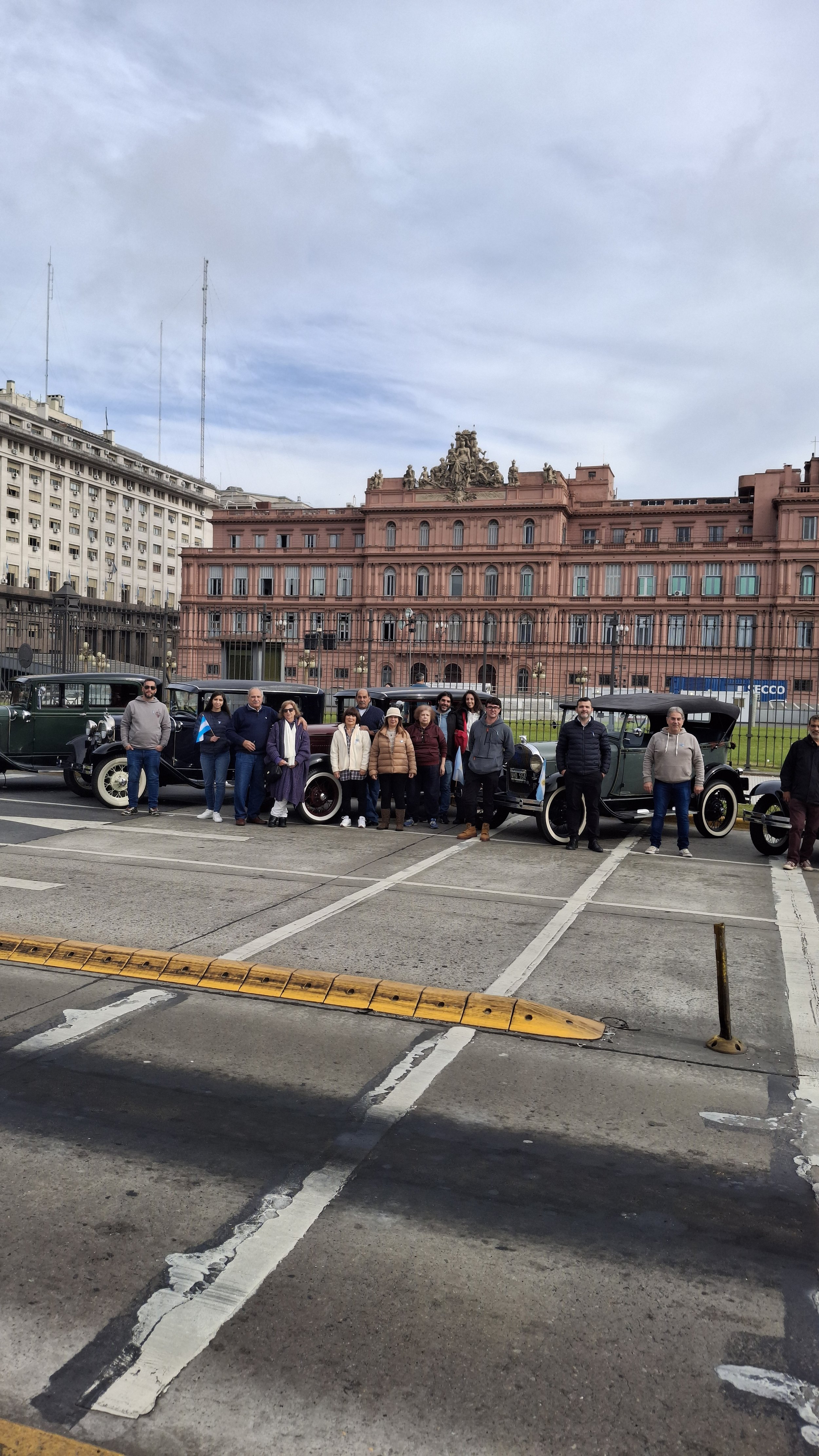 Grupo de personas posando frente a autos antiguos en una plaza, con un edificio histórico de fondo, cielo nublado y línea de demarcación en el suelo.